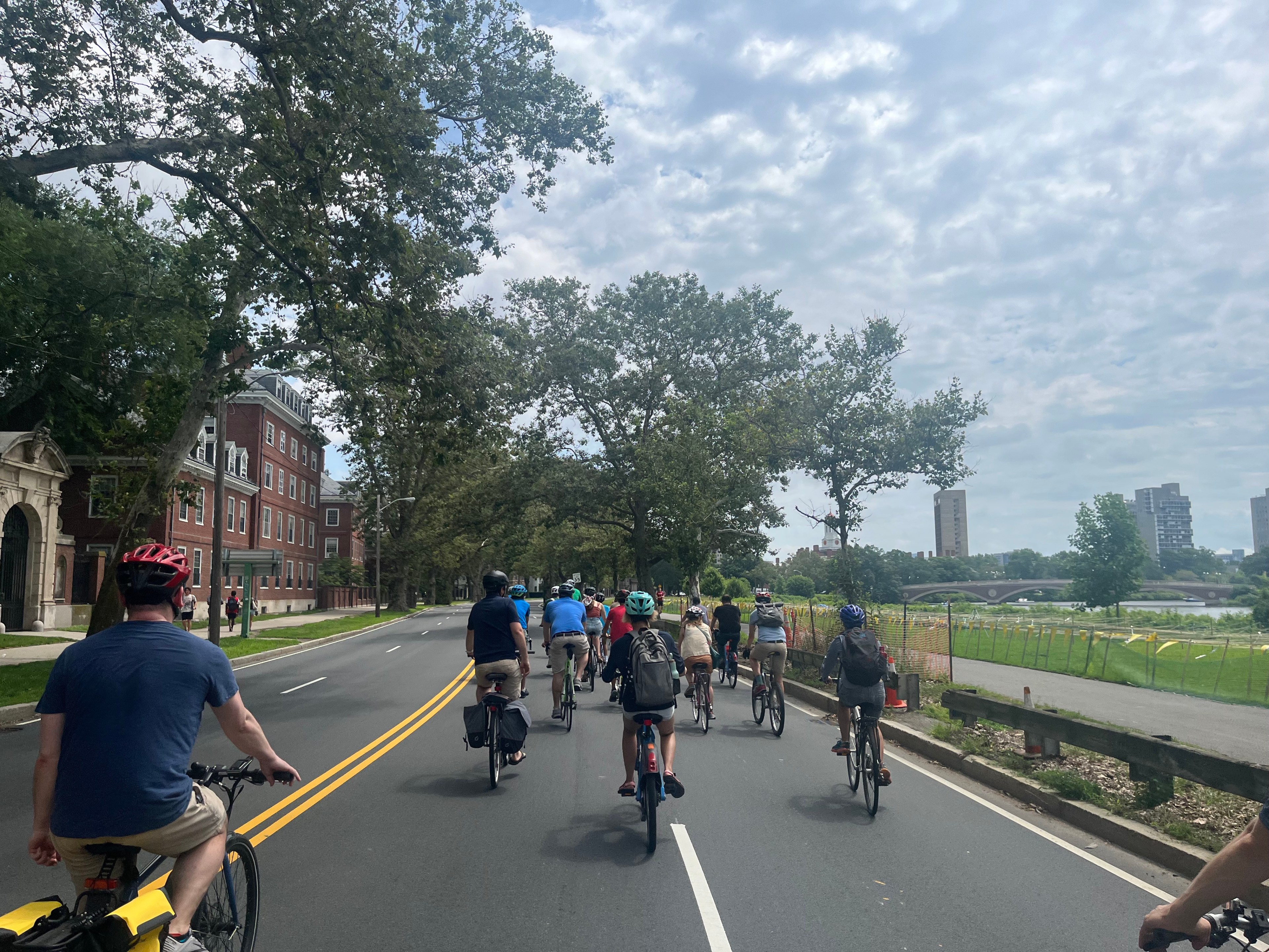 A crowd of people on bikes rides down an empty four-lane highway. On the left side of the roadway are large trees and multi-story brick buildings; on the right is the Charles River with the Boston skyline in the distance.