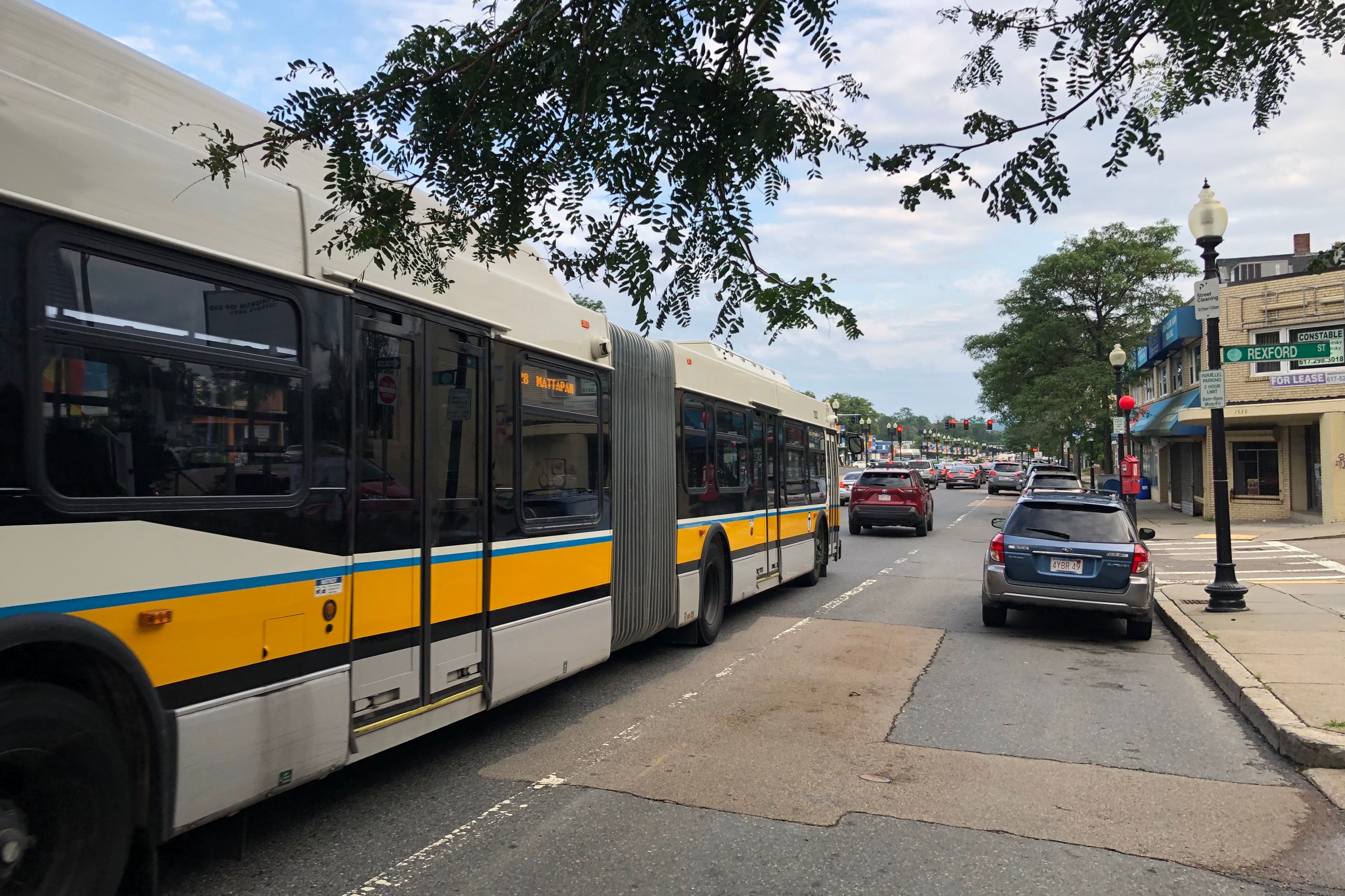 An extra-long articulated bus in the white, yellow, and black MBTA color scheme waits in traffic on a wide multi-lane street with a red light in the distance. An electronic sign in a side window of the bus says "28 MATTAPAN." A street sign on the left says "Rexford Street".
