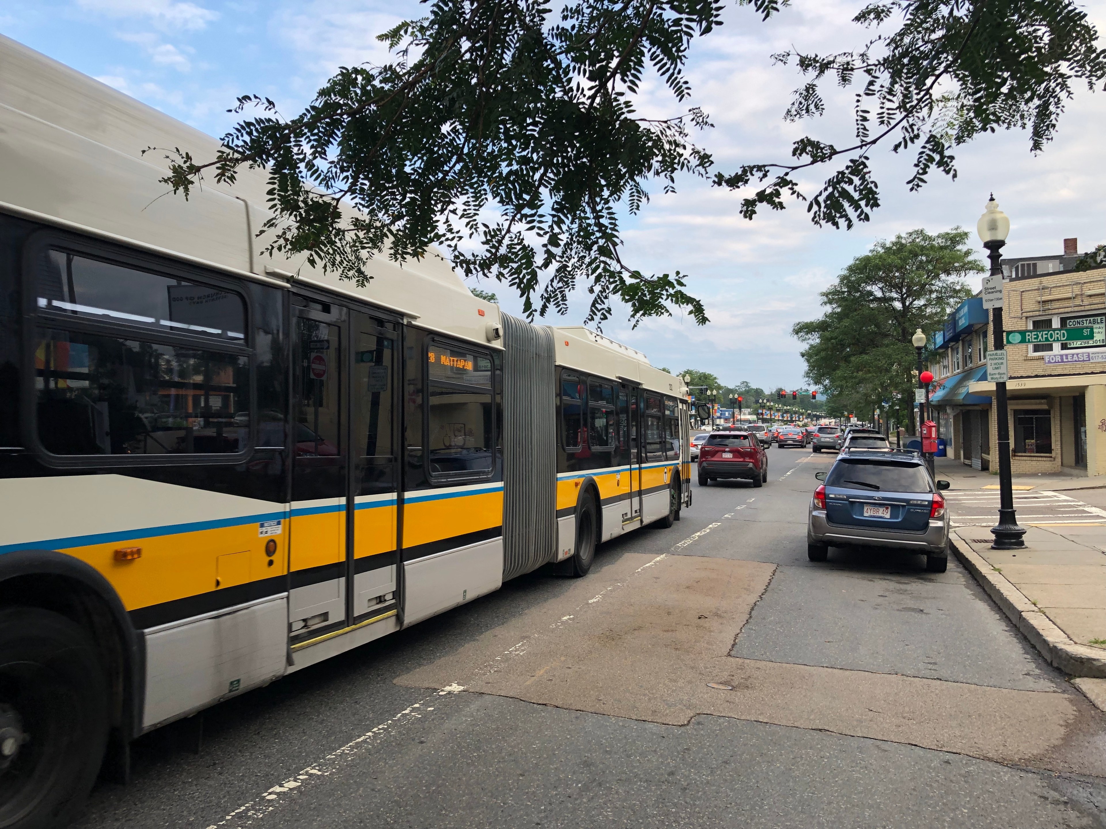An extra-long articulated bus in the white, yellow, and black MBTA color scheme waits in traffic on a wide multi-lane street with a red light in the distance. An electronic sign in a side window of the bus says "28 MATTAPAN." A street sign on the left says "Rexford Street".