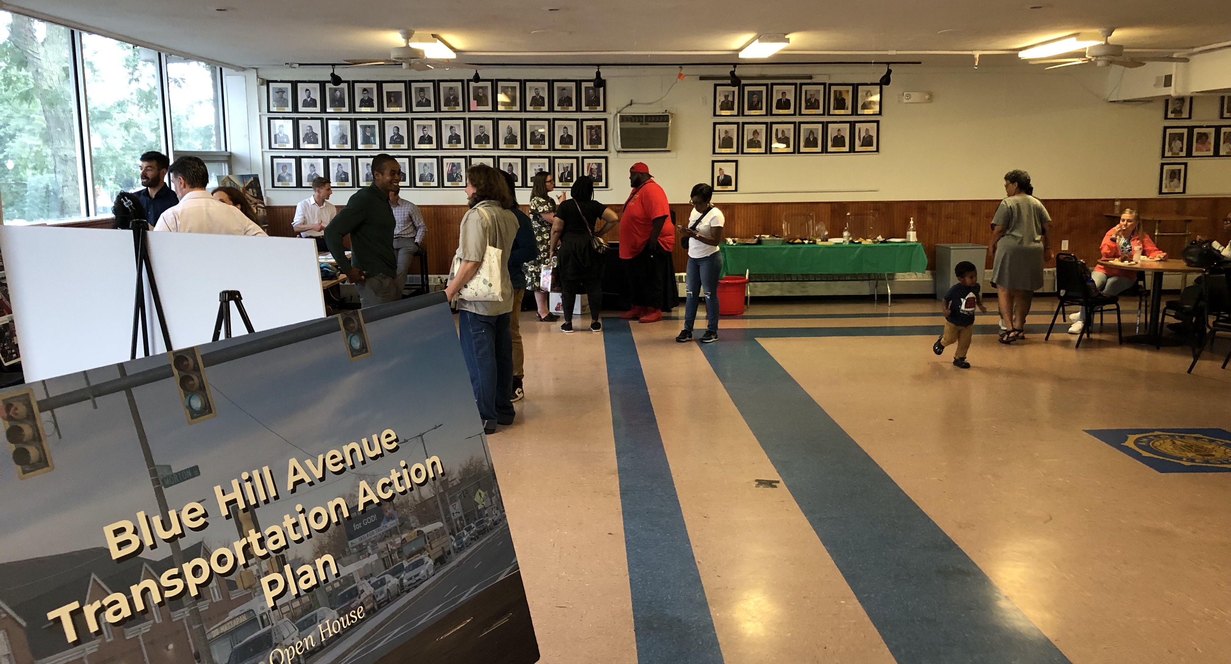 About a dozen people chat in groups in a large function hall. In the foreground at lower left, a poster welcomes attendees with the words "Blue Hill Avenue Transportation Action Plan Open House".