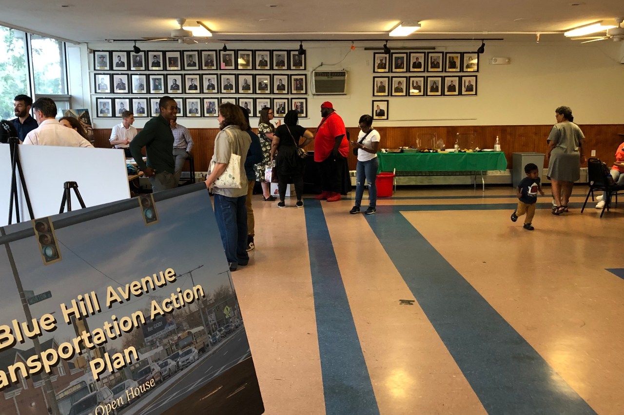 About a dozen people chat in groups in a large function hall. In the foreground at lower left, a poster welcomes attendees with the words "Blue Hill Avenue Transportation Action Plan Open House".