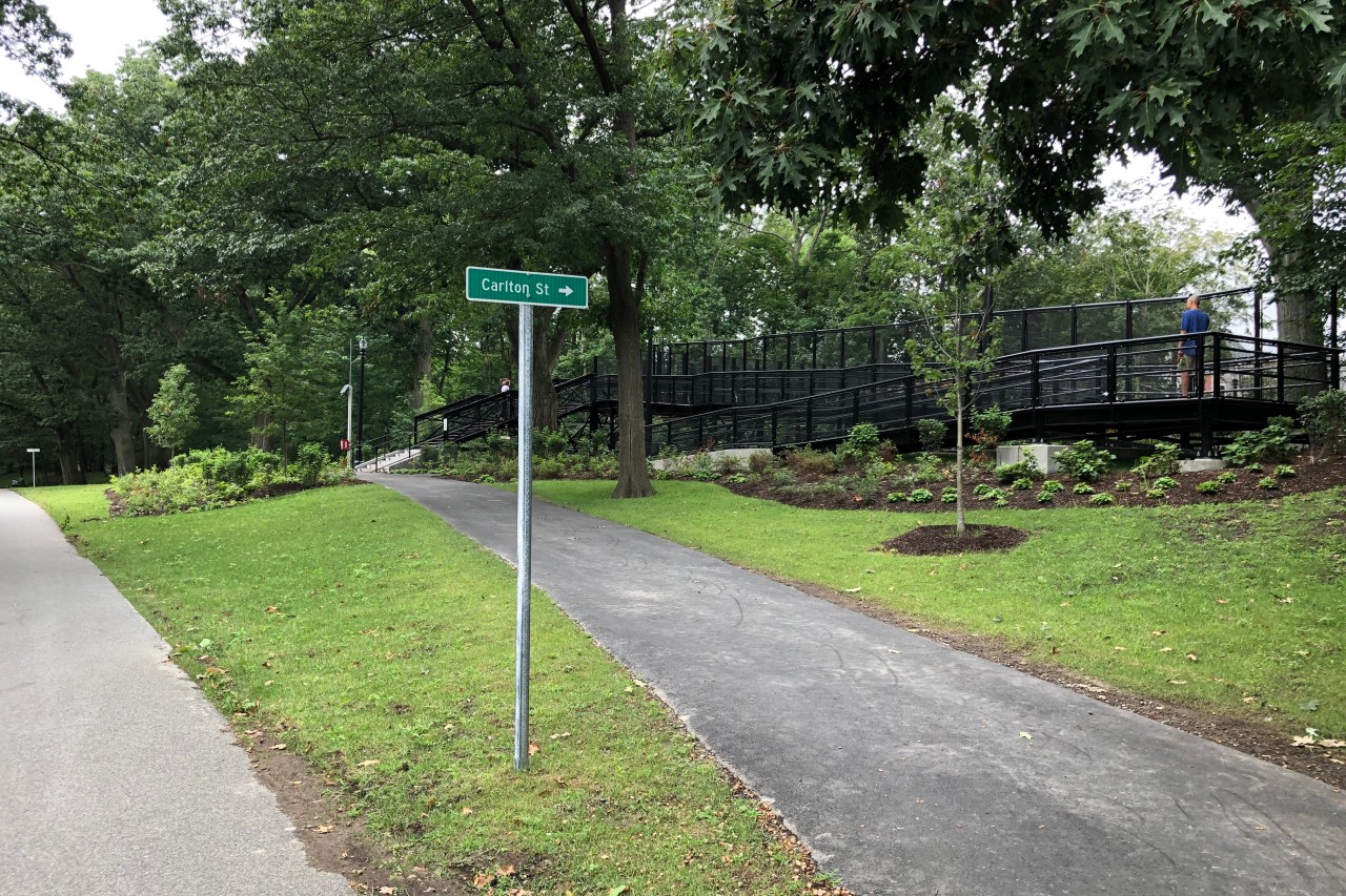 Two paved paths diverge in a green woodland. A green street sign pointing right indicates that the right path leads to Carlton St. At the top of that path, a stairway and a wheelchair ramp made of black metal lead over the Green Line tracks.