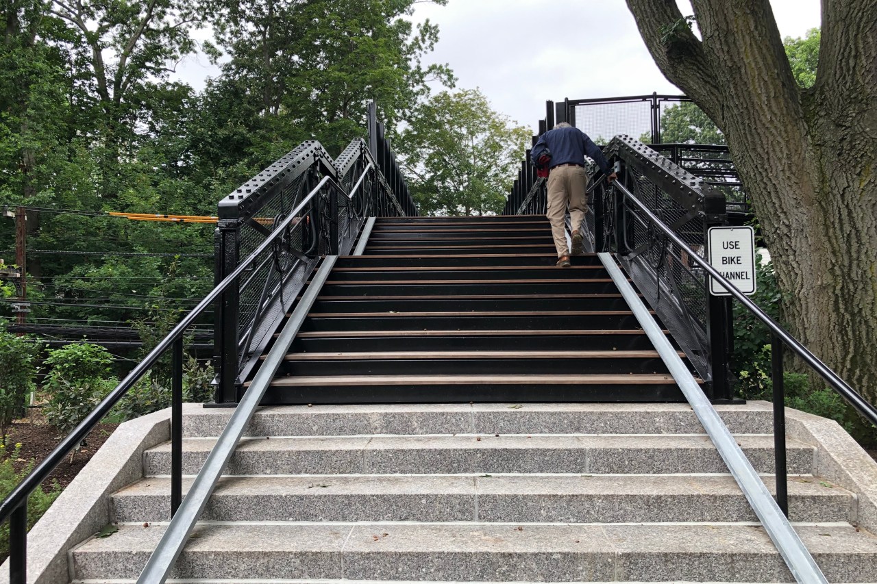 A man in khaki pants climbs a staircase surrounded by trees. At the edges of the stairs, near the base of the railings, are two metal gutters, roughly 6 inches wide, which are ramps designed to let people roll their bikes up and down the steps. A sign to the right reads "USE BIKE CHANNEL"