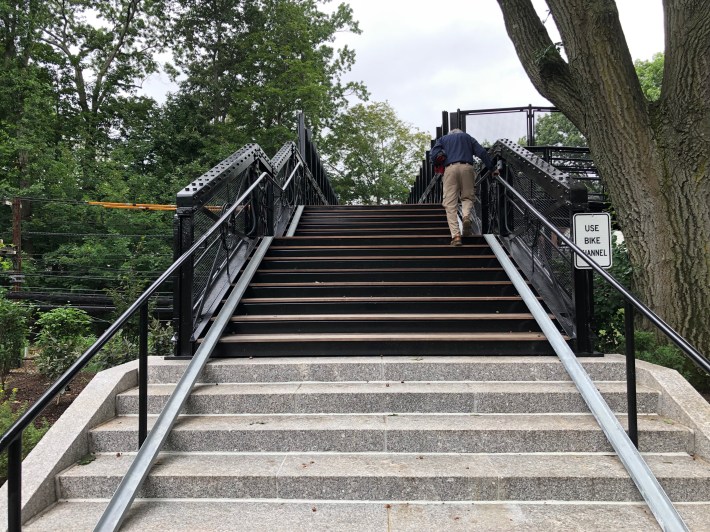 A man in khaki pants climbs a staircase surrounded by trees. At the edges of the stairs, near the base of the railings, are two metal gutters, roughly 6 inches wide, which are ramps designed to let people roll their bikes up and down the steps. A sign to the right reads "USE BIKE CHANNEL"