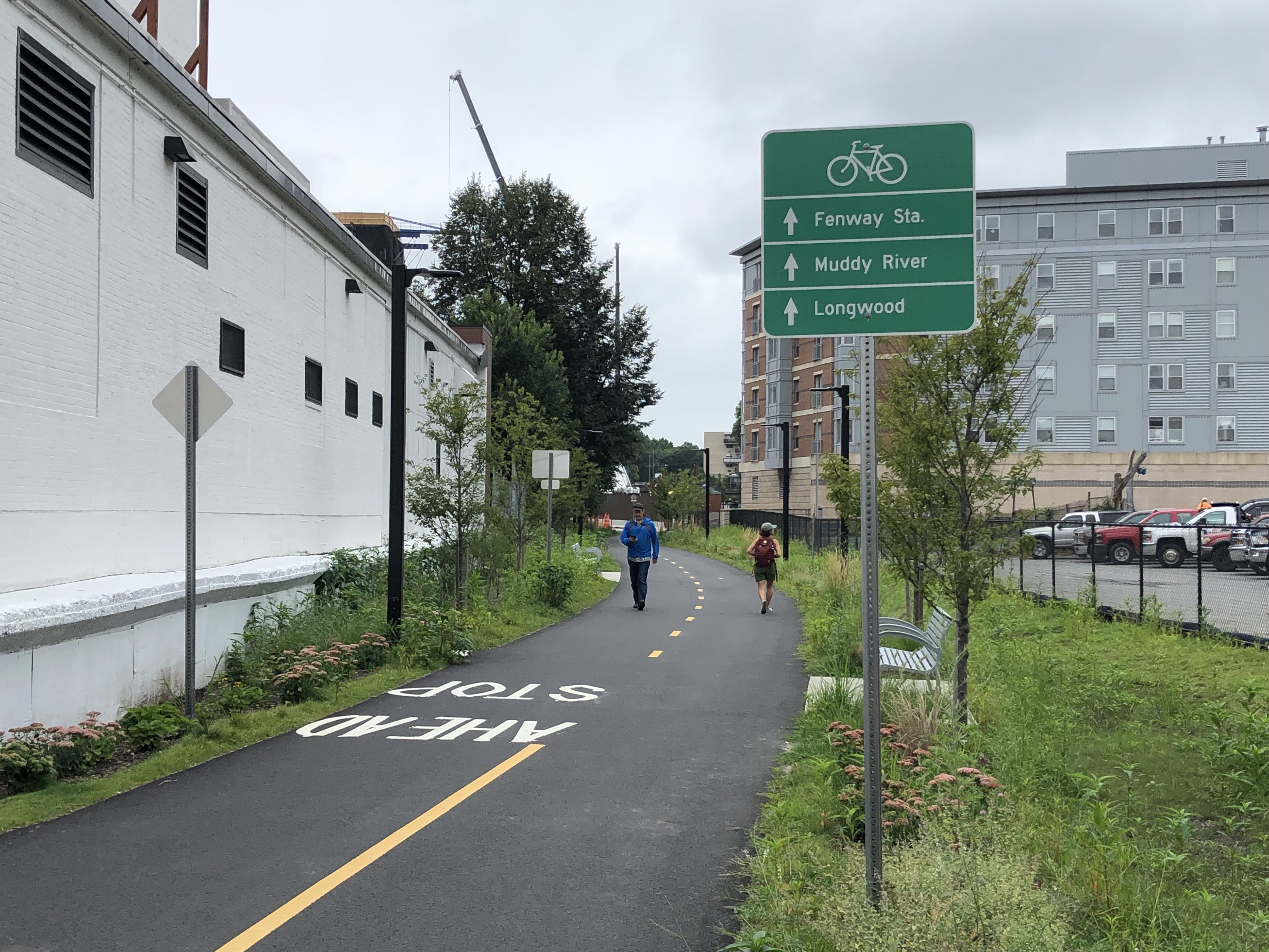 A paved asphalt bike and pedestrian path winds behind a single-story cinder block building on the left and a parking lot on the right. A green wayfinding sign shows three destinations, listed next to arrows that point straight down the path: Fenway Sta., Muddy River, and Longwood. In the middle distance two people walking are passing each other on the path, and further on in the distance is a crane at a construction site behind a multi-story apartment building.