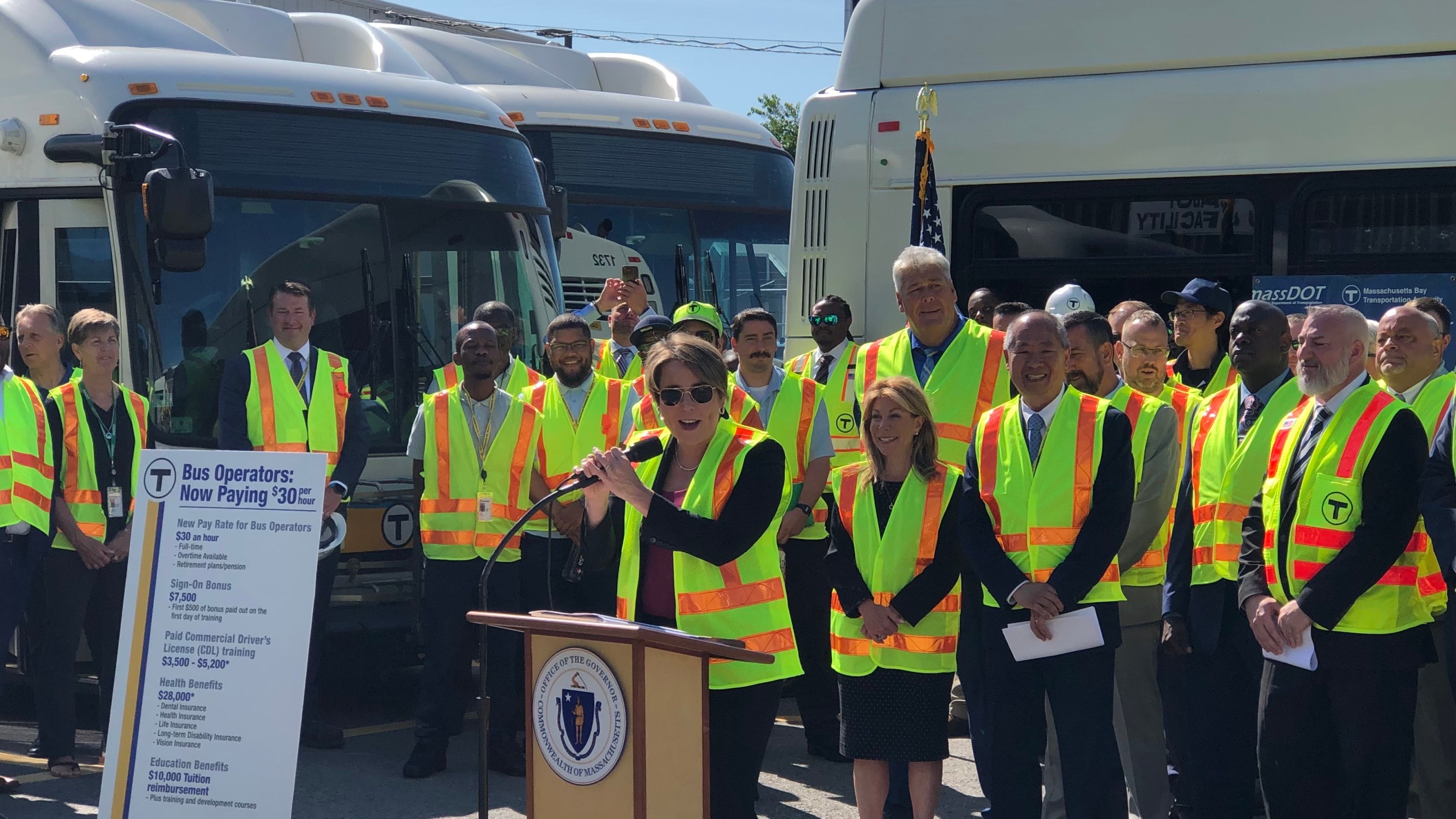 A crowd of people in yellow flourescent safety vests stands behind a podium where the Massachusetts Governor is speaking. To her left is a poster that outlines the new wages and benefits for newly-hired bus drivers under the terms of a new labor deal, with a $30 per hour wage in bold lettering at the top.
