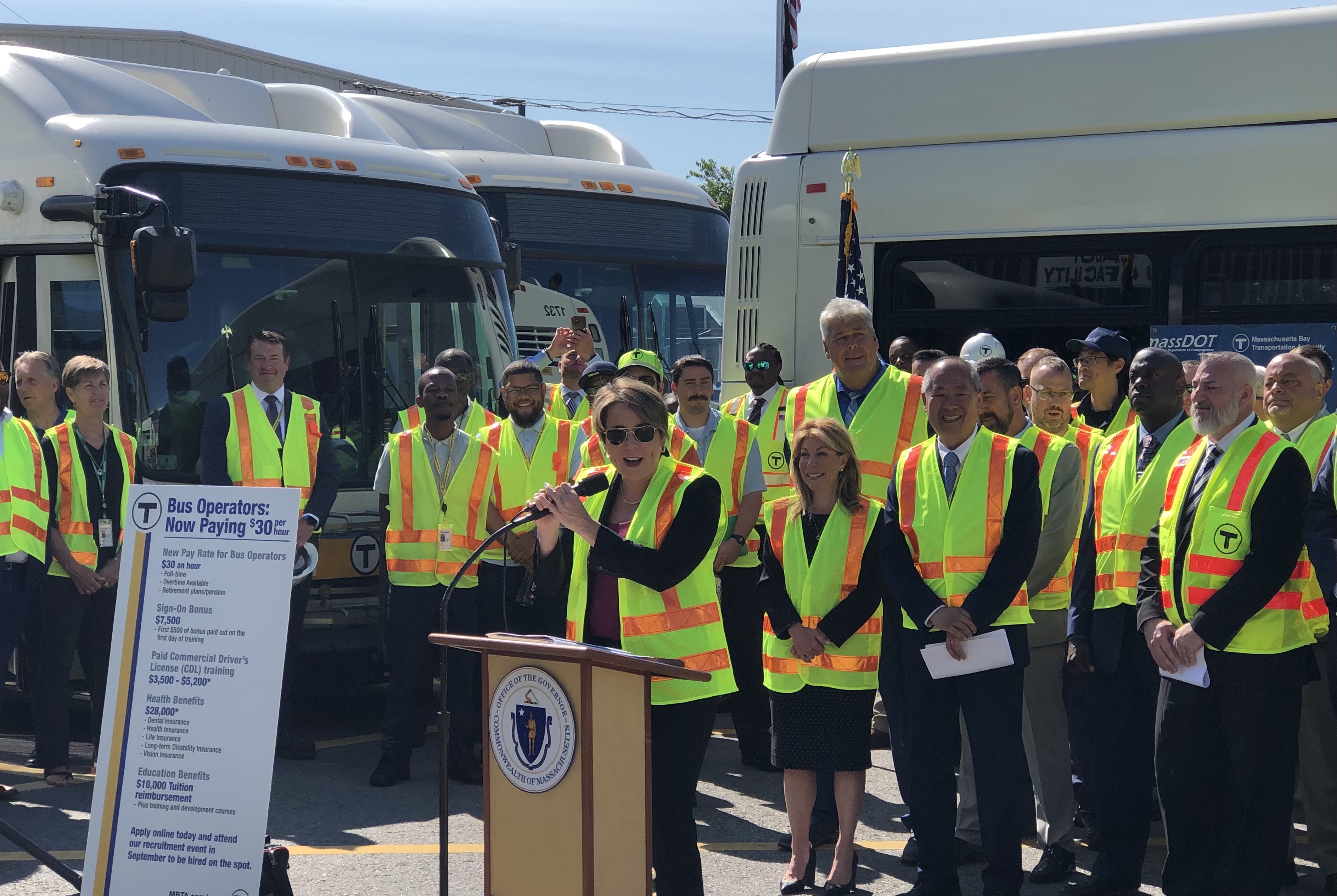 A crowd of people in yellow flourescent safety vests stands behind a podium where the Massachusetts Governor is speaking. To her left is a poster that outlines the new wages and benefits for newly-hired bus drivers under the terms of a new labor deal, with a $30 per hour wage in bold lettering at the top.