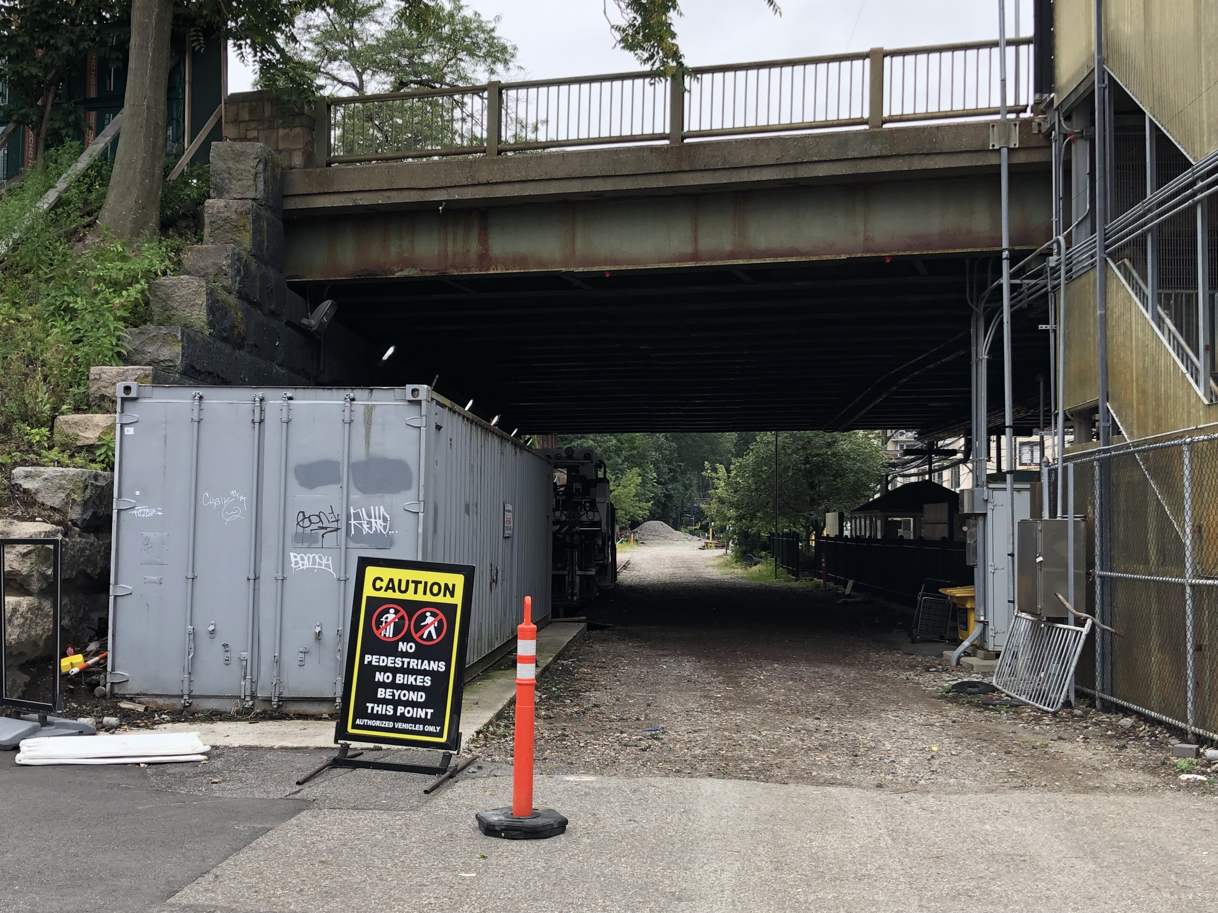A gravel area underneath a roadway bridge. In the distance on the other side of the bridge trees are visible. To the right a staircase leads from the ground level to the bridge. In the foreground a sign says "Caution no pedestrians no bikes beyond this point - authorized vehicles only"
