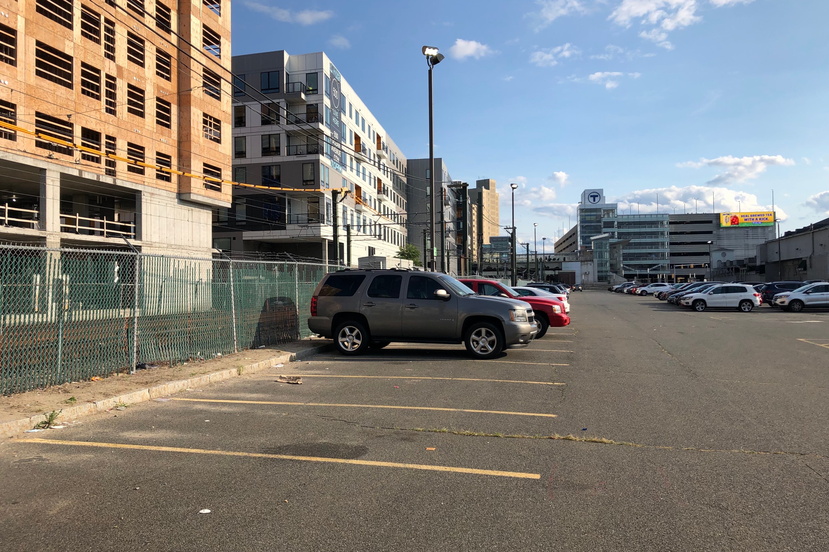 A half-empty parking lot next to several mid-rise apartment buildings, one of which (on the left edge of the photo) is still under construction. In the distance is a large parking garage with the MBTA logo on top. Some railroad tracks – the MBTA Blue Line – are visible through a chain-link fence at left, between the apartments and the parking lot.