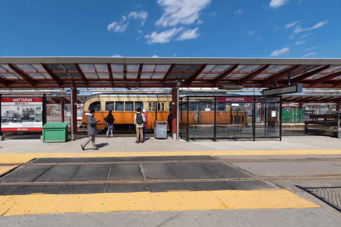 Riders wait on a platform under a canopy with an orange streetcar waiting on the other side of the platform. Signage on the platform indicates that this is the Mattapan station.