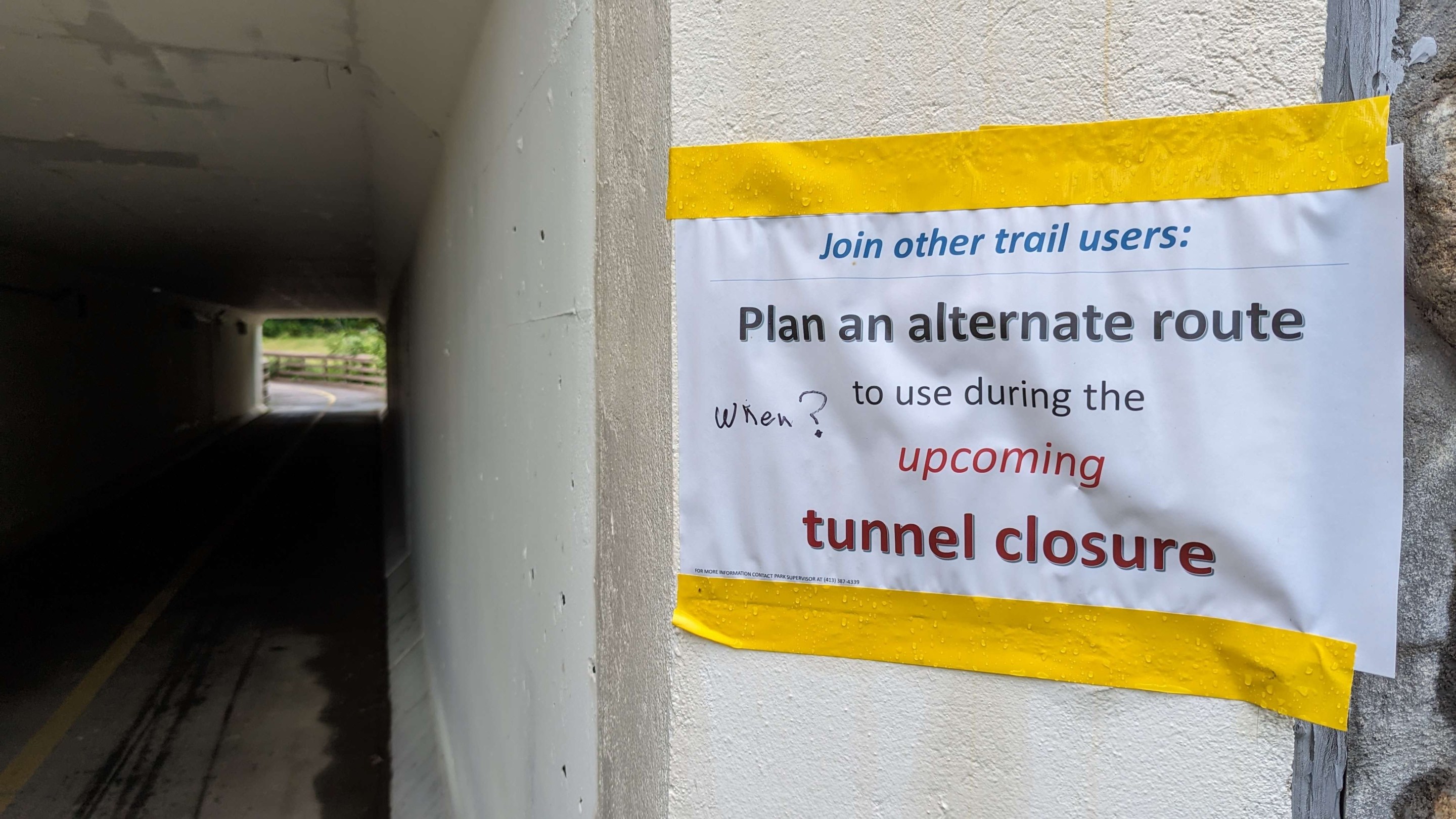 A homemade sign printed on 8.5"x11" paper is haphazardly taped to the entrance to a pedestrian tunnel. The sign uses different fonts for each line of text. The top line reads (in blue, italicized text): "Join other trail users:" the next line, in black, larger text: "Plan an alternate route", the next line, smaller black text: "to use during the" (in red, larger italicized text) "upcoming" (final line, red bolder text) "tunnel closure". On the left side of the paper, someone has hand-written the word "When?" with a black marker. Fine print at the bottom of the sign says "FOR MORE INFORMATION CONTACT PARK SUPERVISOR AT (413) 387-4339."