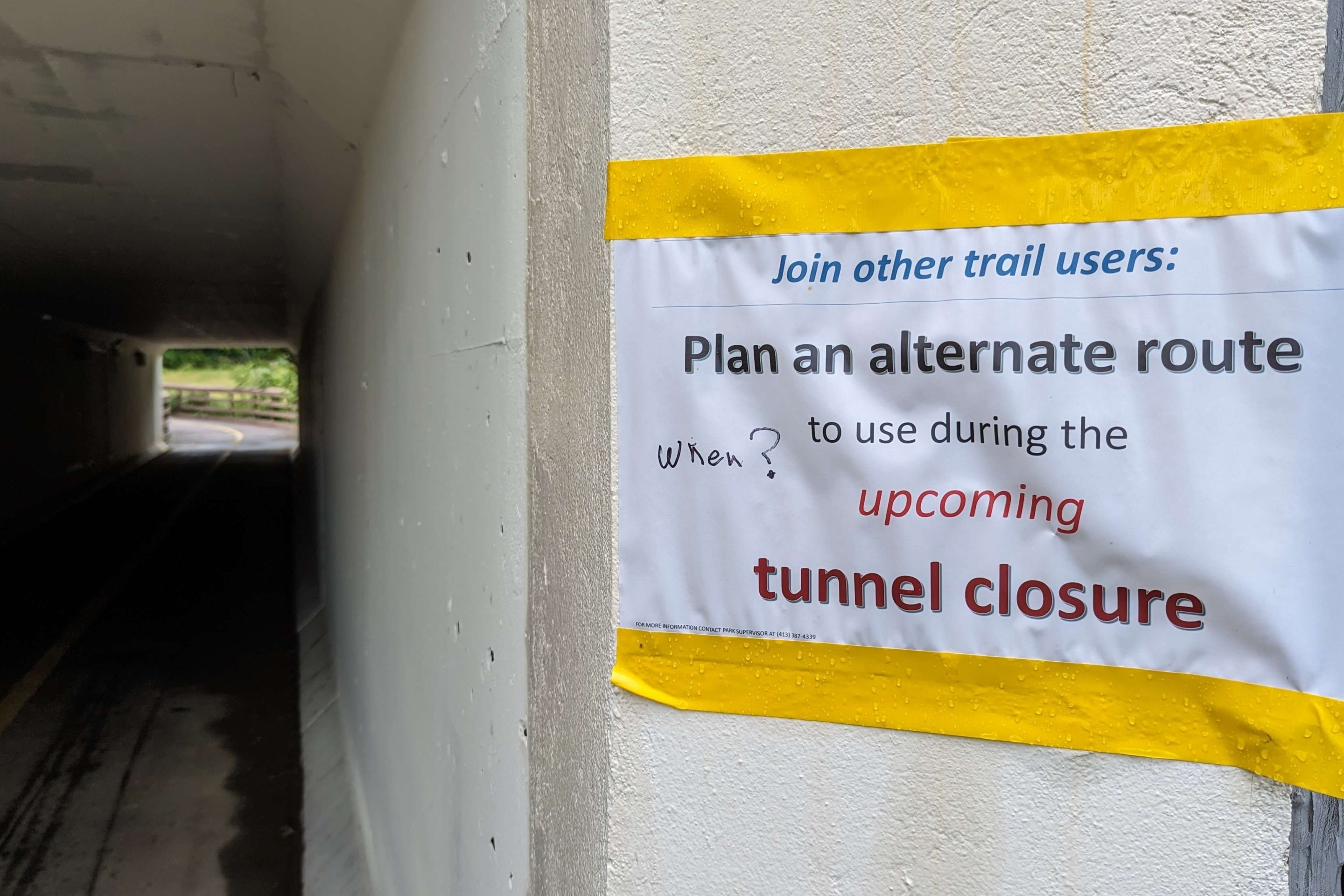 A homemade sign printed on 8.5"x11" paper is haphazardly taped to the entrance to a pedestrian tunnel. The sign uses different fonts for each line of text. The top line reads (in blue, italicized text): "Join other trail users:" the next line, in black, larger text: "Plan an alternate route", the next line, smaller black text: "to use during the" (in red, larger italicized text) "upcoming" (final line, red bolder text) "tunnel closure". On the left side of the paper, someone has hand-written the word "When?" with a black marker. Fine print at the bottom of the sign says "FOR MORE INFORMATION CONTACT PARK SUPERVISOR AT (413) 387-4339."