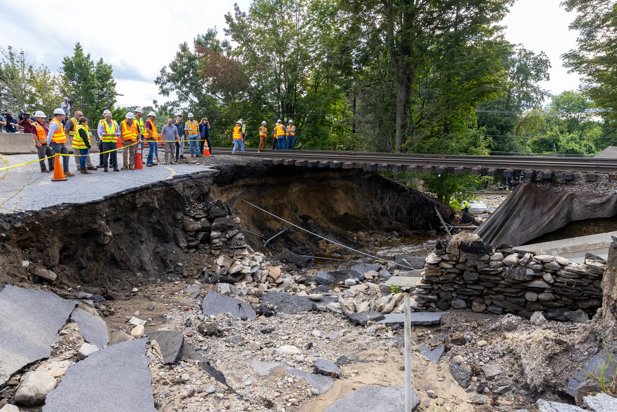 A group of people in construction vests stands on asphalt next to a large, washed-out gully that has destroyed a roadway and undermined a railroad. To their right, steel rails and their wooden ties are suspended in midair above the washout.
