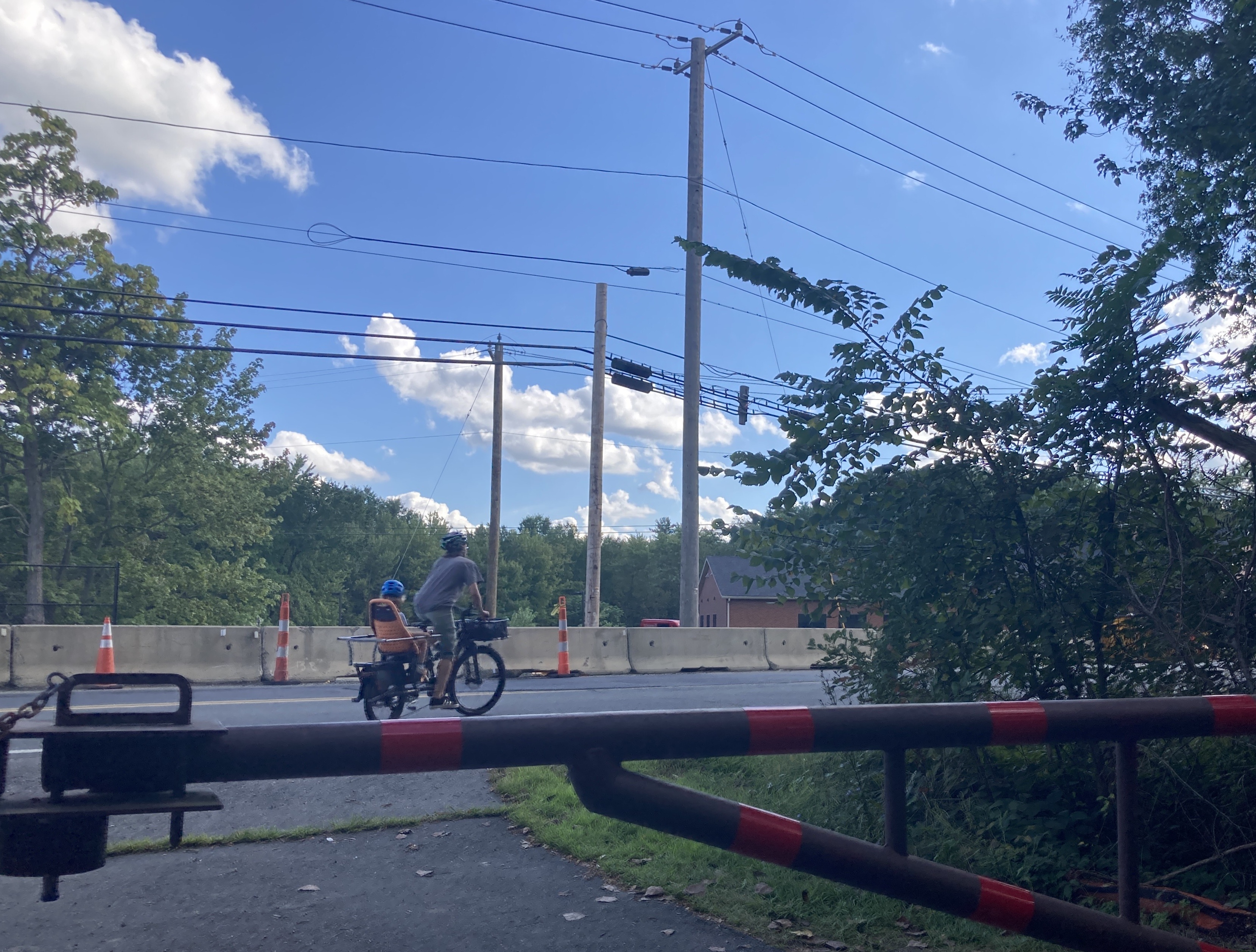 A man and child riding. a bike onto a 2-lane roadway. A row of jersey barriers blocks the opposite side of the street. Beyond are some power lines and trees, and in the foreground is a gate.