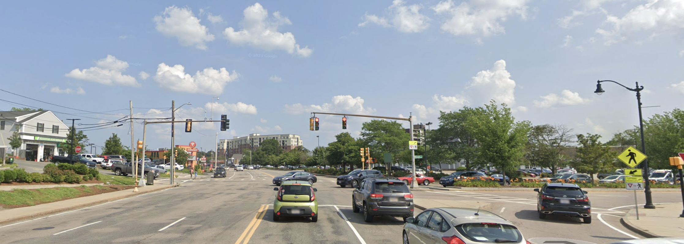A Google Street View image of a wide intersection. In the foreground are five lanes of traffic with cars waiting at a red light. To the right is an intersecting multi-lane roadway. In the distance is a multi-story apartment building.