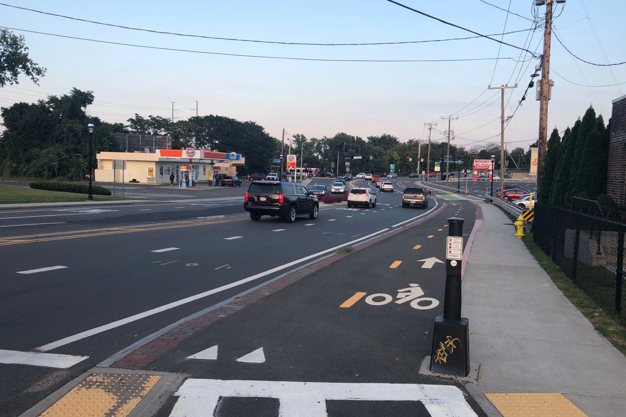 A paved bike path runs alongside a wide multi-lane road (at left) and a sidewalk (at right) with a striped crosswalk in the foreground. To the left of the roadway is a gas station and to the right is a parking lot.