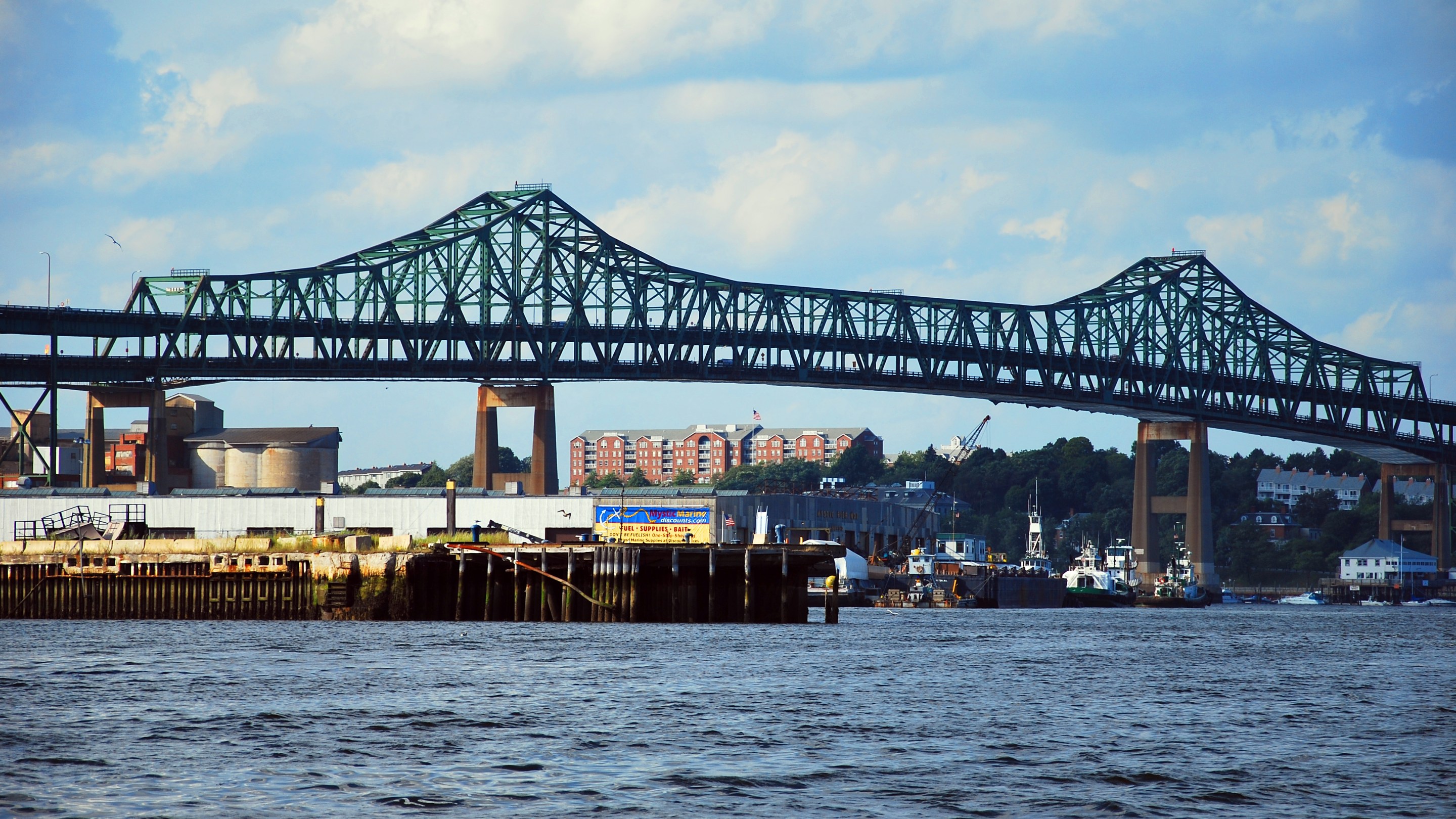A large bridge painted green spans a choppy body of water above some tugboats and wharves.