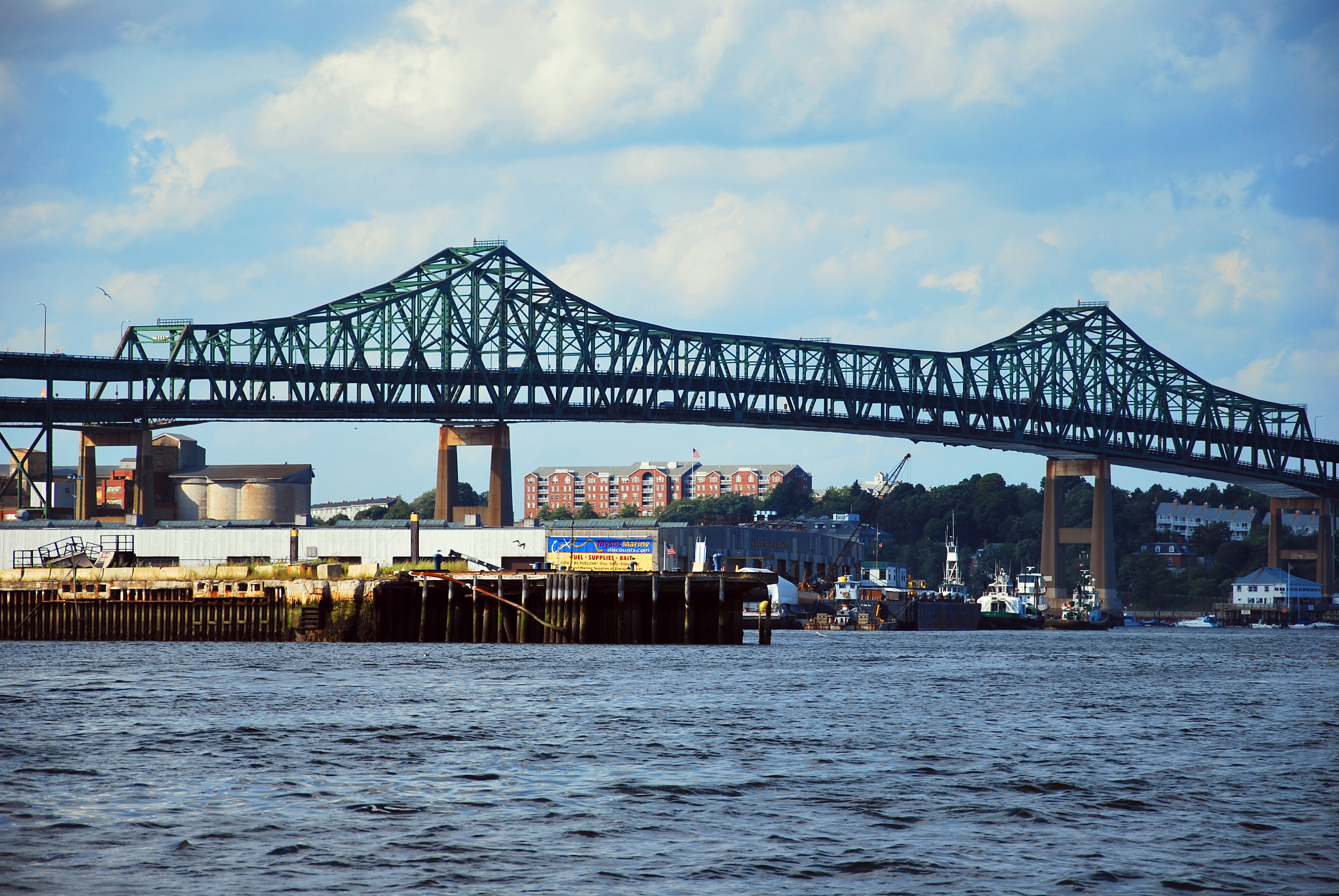 A large bridge painted green spans a choppy body of water above some tugboats and wharves.