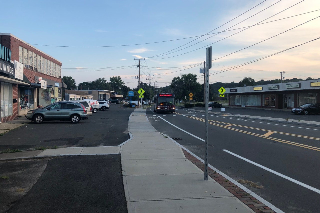 A PVTA bus approaches along a two-way street lined with strip malls and parking lots. The street includes new sidewalks and painted bike lanes and the strip malls on either side are full of small businesses.