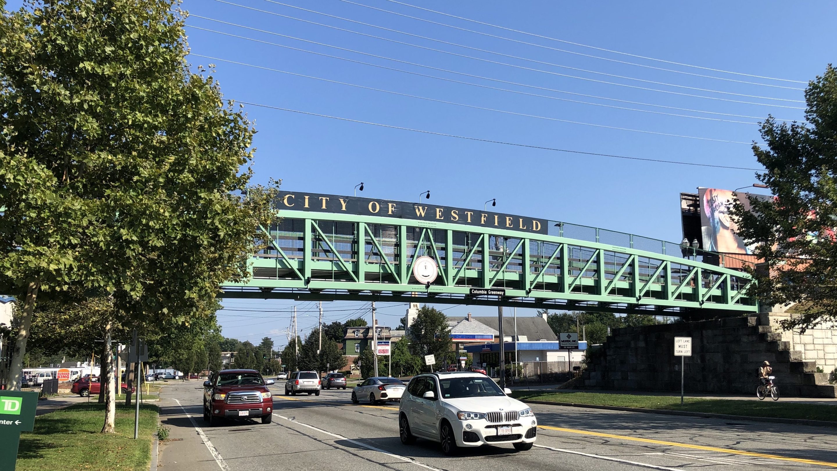 A green truss bridge spans a wide five-lane roadway with two leafy trees on either side on a clear sunny day. A sign along the top railing of the bridge says "CITY OF WESTFIELD" while a smaller sign on the bottom reads "Columbia Greenway". In between the two signs is a round city seal in the center of the bridge.