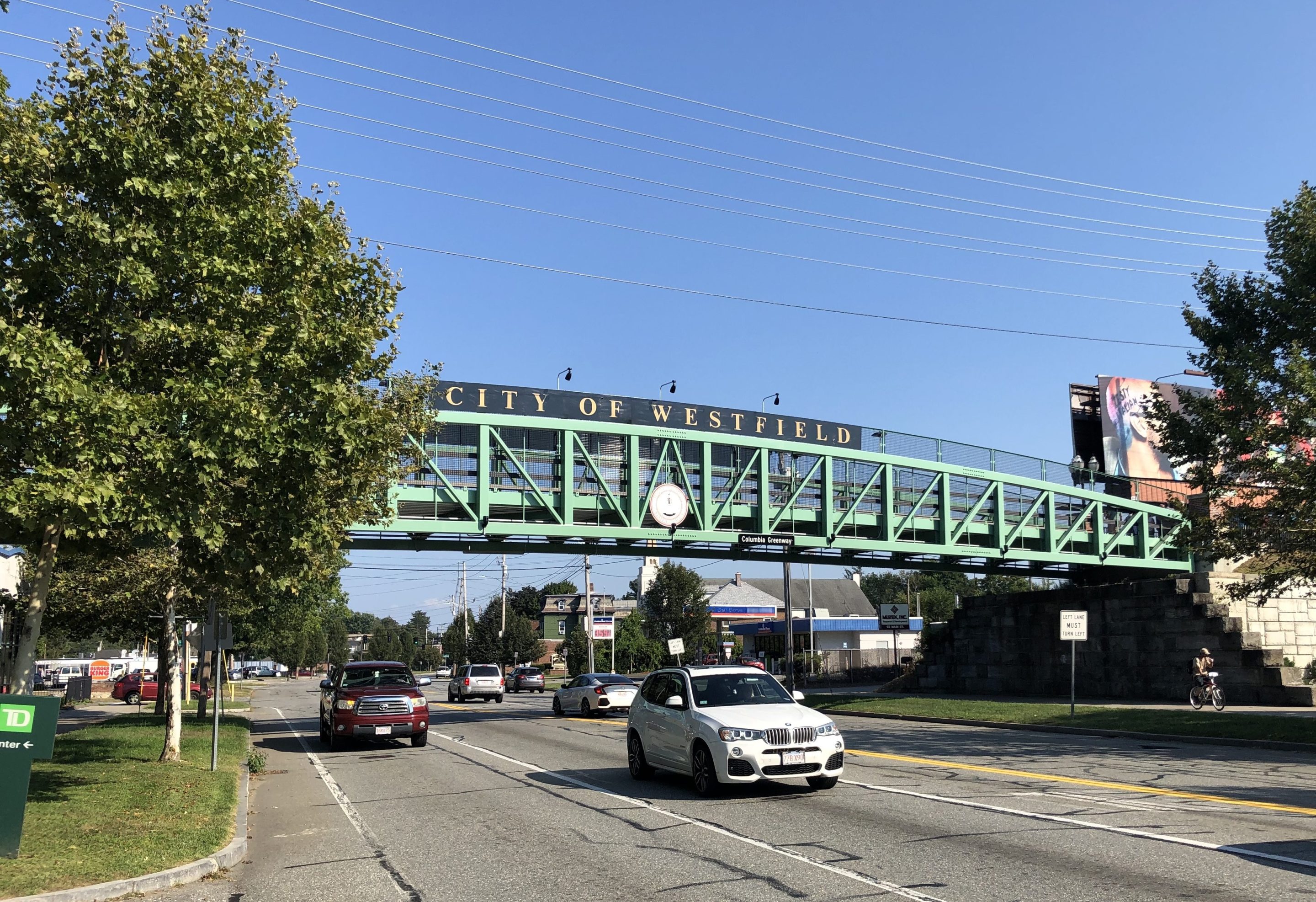 A green truss bridge spans a wide five-lane roadway with two leafy trees on either side on a clear sunny day. A sign along the top railing of the bridge says "CITY OF WESTFIELD" while a smaller sign on the bottom reads "Columbia Greenway". In between the two signs is a round city seal in the center of the bridge.