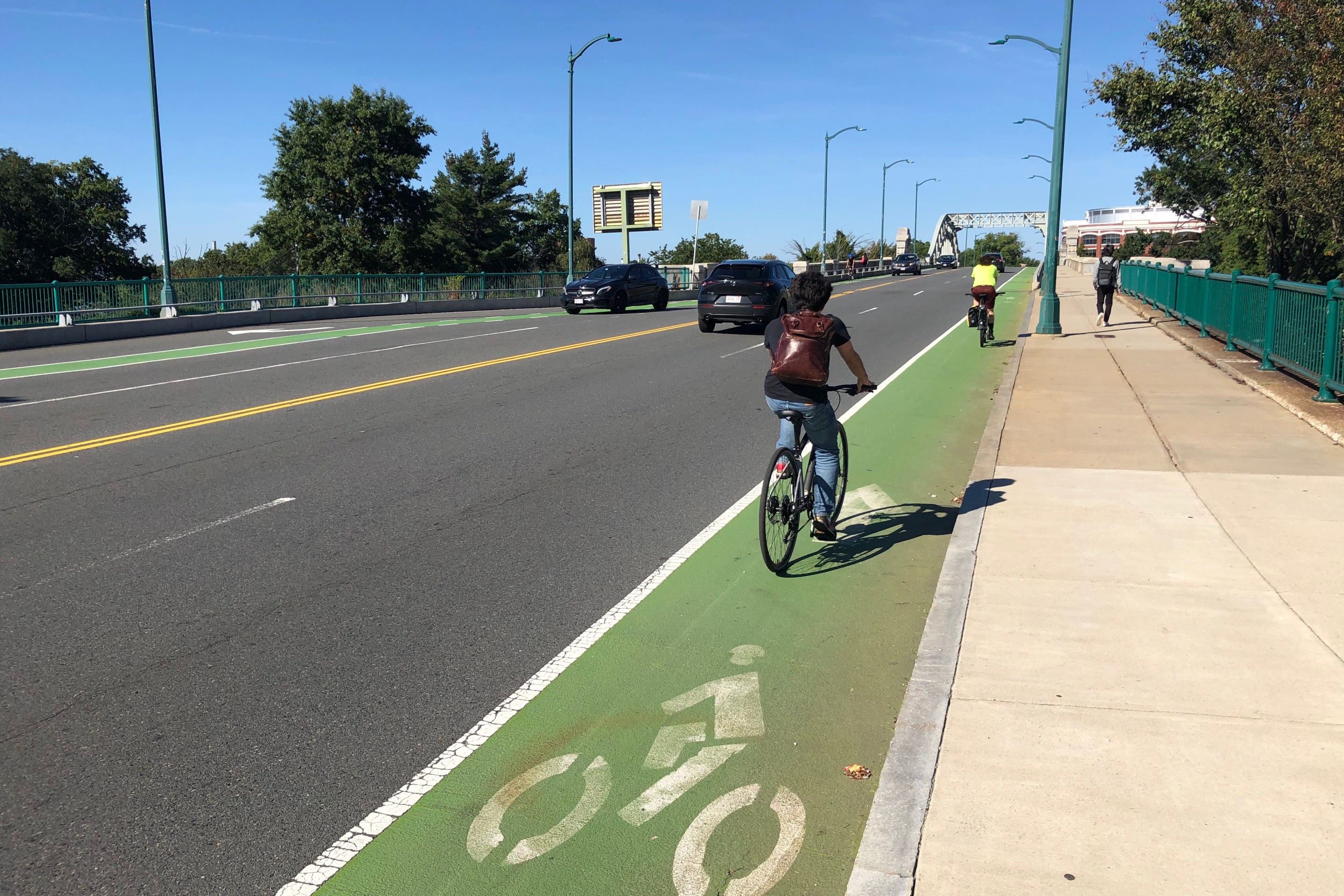 Two people on bikes ride on a green-painted bike lane towards an arched bridge on the horizon. Next to them are four lanes for motor vehicles being used by two cars, and a wide sidewalk on their right.