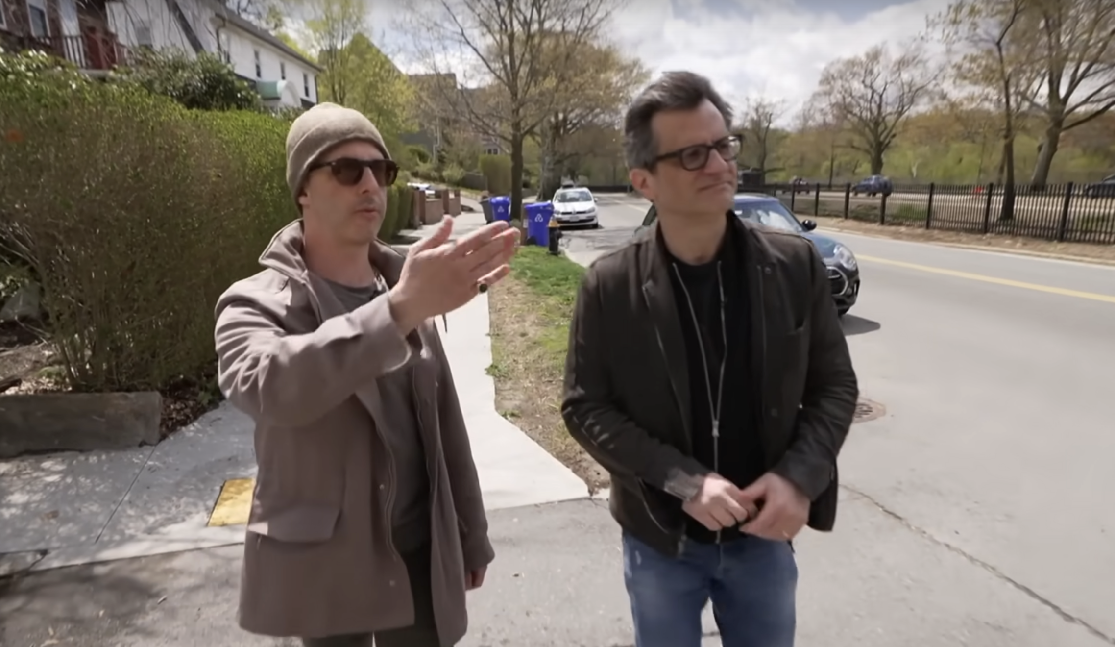 Two men in coats walk across a small street next to a much larger multi-lane roadway. The man on the left, wearing sunglasses, is Jeremy Strong. He is gesturing to the large road to his left. The man on the right, reporter Ben Mankiewicz, is looking in the direction of Strong's gesture.