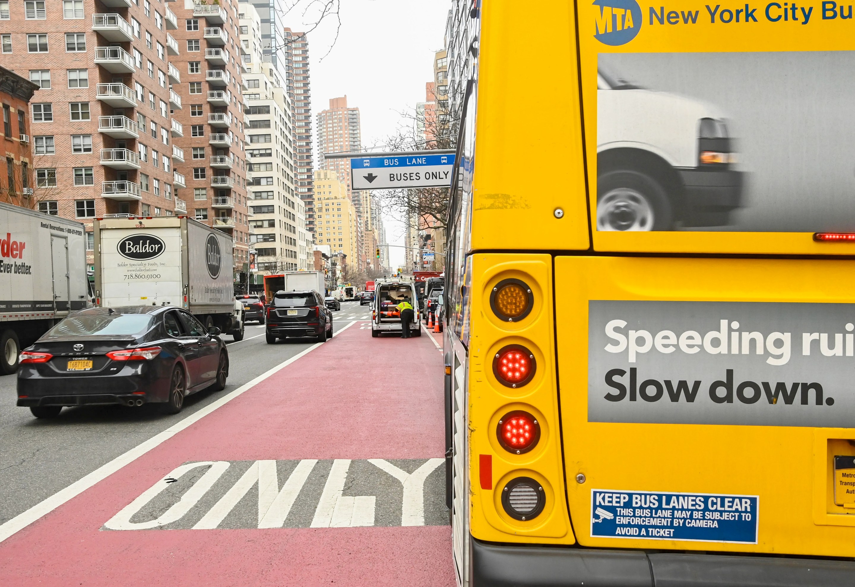 A view of a New York city street with a red "BUS ONLY" lane in the middle of the image, and the yellow rear end of an MTA city bus at right. The rear of the bus includes a blue sticker that says "KEEP BUS LANES CLEAR" and "This bus lane may be subject to enforcement by camera - avoid a ticket"
