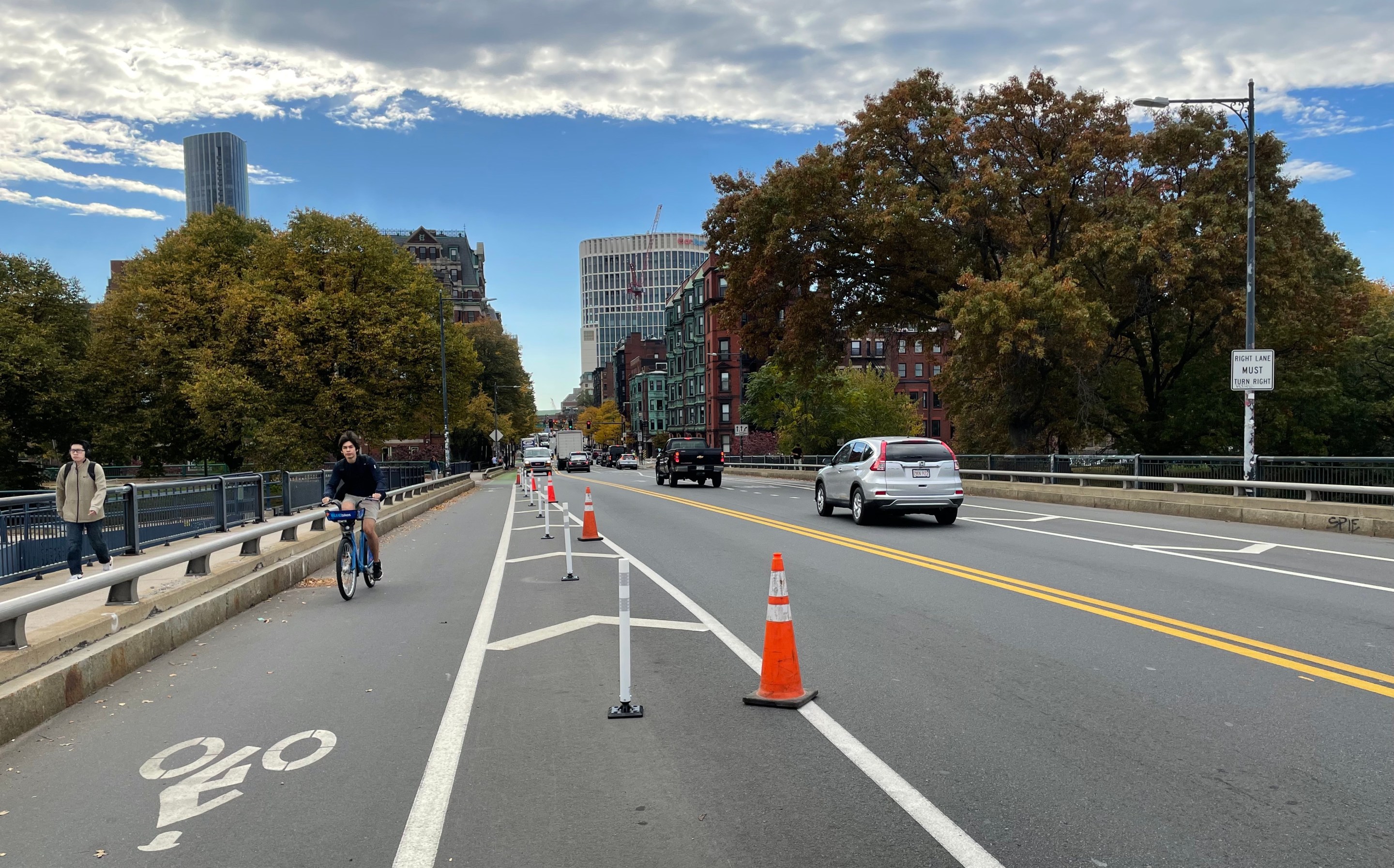 A photograph of a bridge with a bike lane at left, and a man riding a bike toward the camera, and two car lanes to the right. A column of white flexible-post bollards with a handful of orange construction cones occupies the buffer zone between the bike lane and the car lanes. In the distance, at the end of the bridge, are some trees and the tall buildings of the Boston skyline.