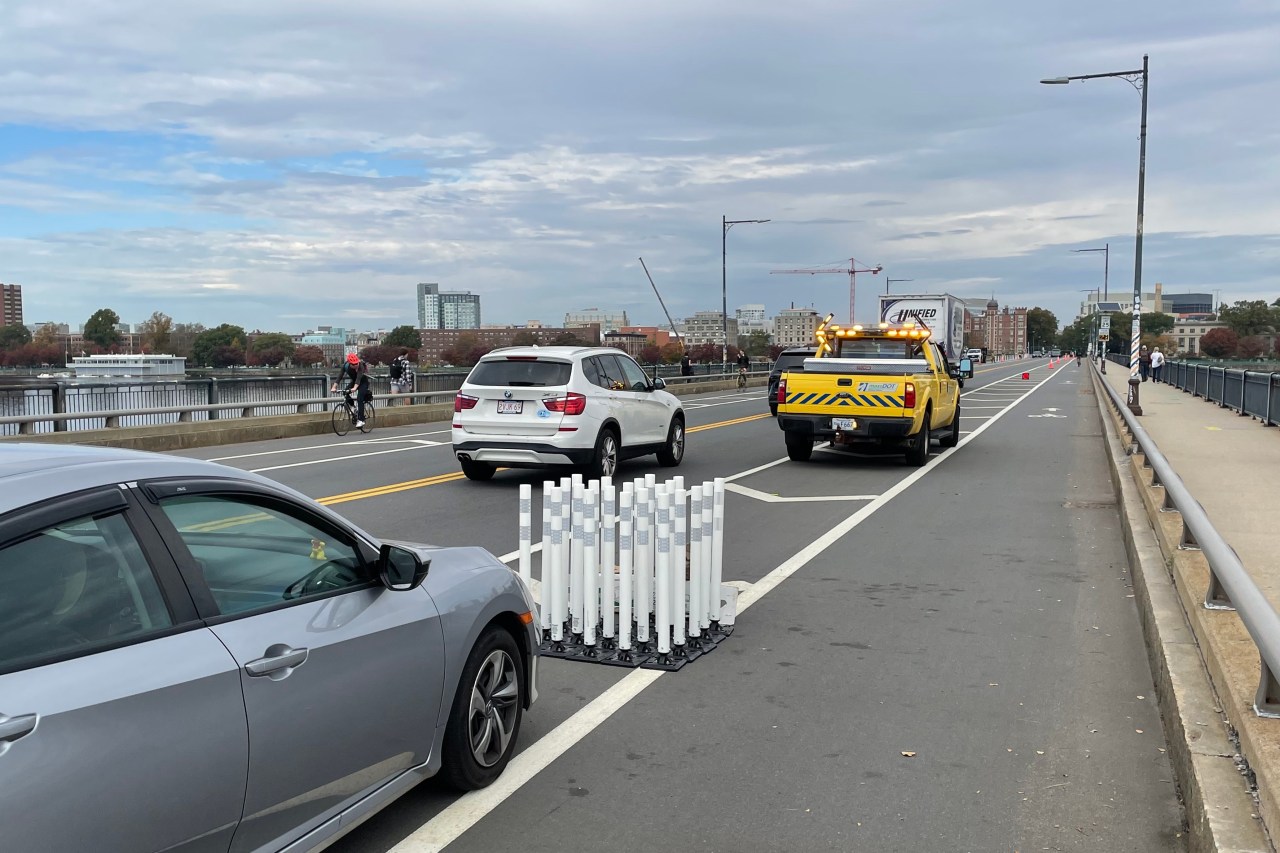A photograph of a bridge with a bike lane and wide sidewalk at right, and two MassDOT vehicles in the buffer zone between the car lane and the bike lane. A cluster of white flexible-post bollards is bundled between the two vehicles. In the distance, at the end of the bridge, is the Cambridge skyline.