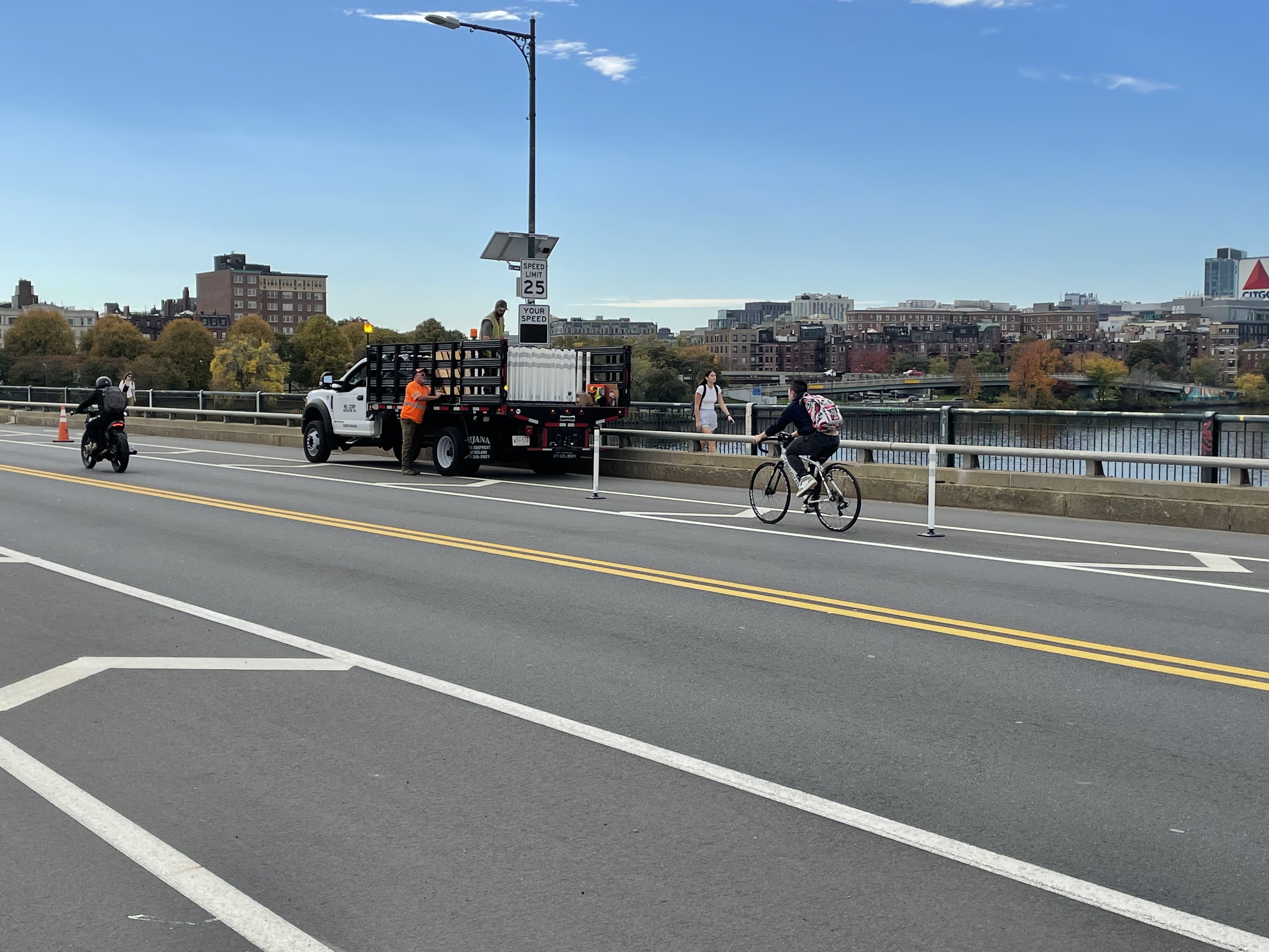 A bicyclist rides past a flatbed truck where two workers are unloading white flexible-post bollards onto the bridge deck.