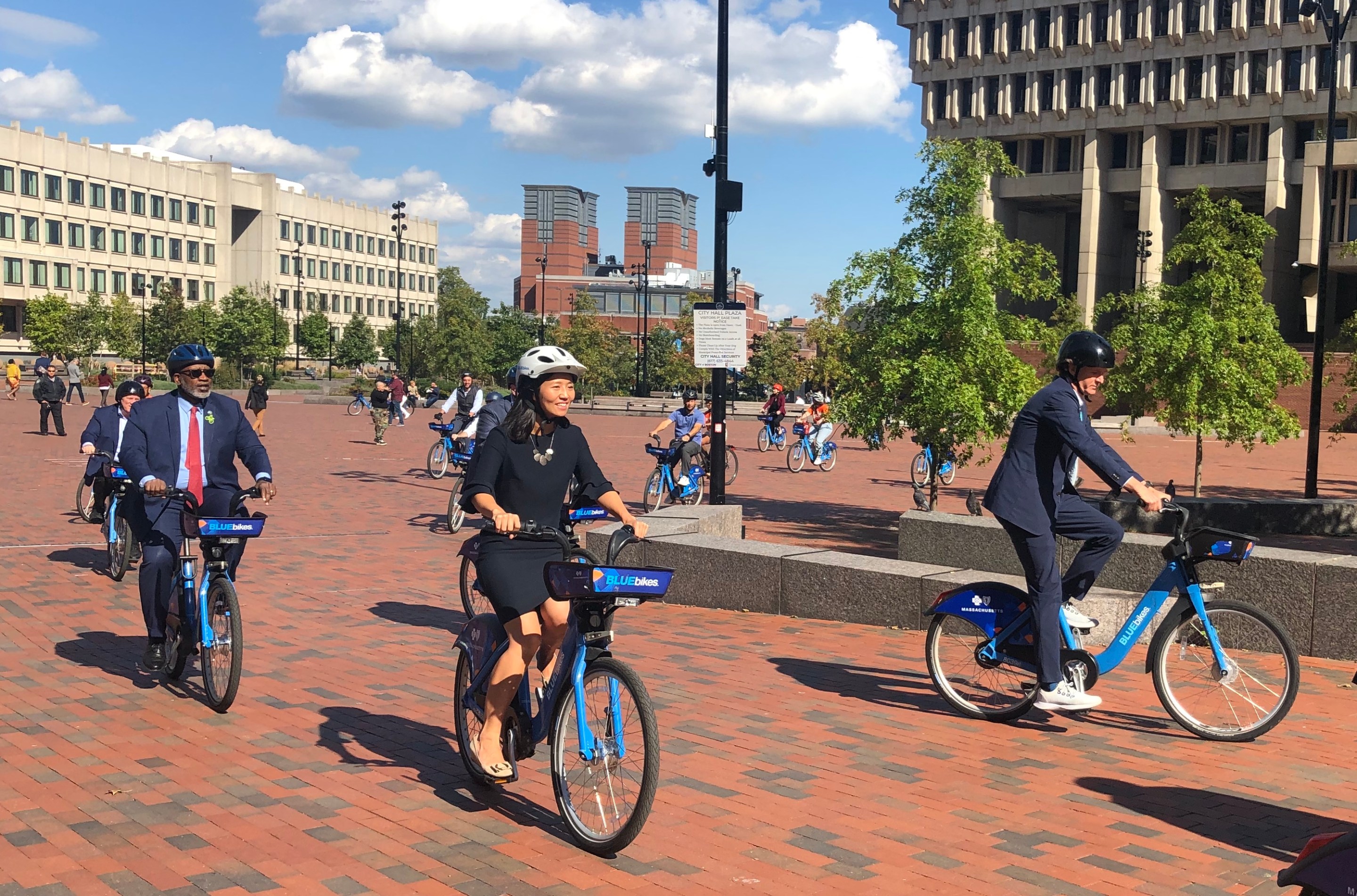 Several people in business suits ride Bluebikes across the bricks of City Hall Plaza under blue skies. Boston's concrete City Hall is visible in the right background.