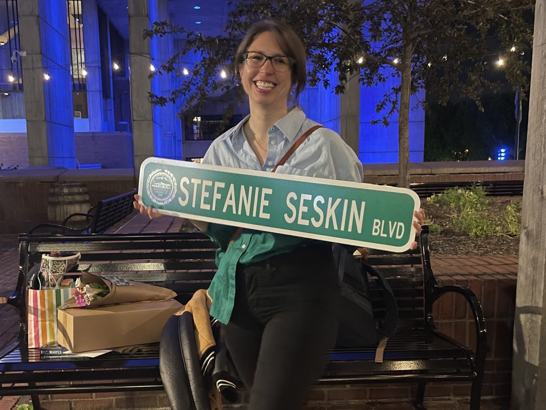A nighttime photograph of a smiling woman wearing glasses poses holding a green street sign that reads "STEFANIE SESKIN BLVD". Behind her, Boston City Hall is lit up in blue lighting.