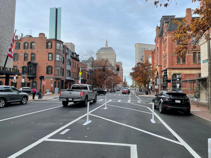 A city street lined with historic multi-story brick rowhouse buildings with the glass Hancock Tower skyscraper rising above them in the background. In the foreground, a broad area of pavement with hatched white markings and flexible-post bollards delineates a wide buffer zone between a bike lane (right) and moving traffic (at left); however, there is a black sedan parked in the bike lane.