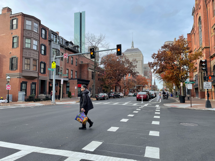 A city street lined with historic multi-story brick rowhouse buildings with the glass Hancock Tower skyscraper rising above them in the background. In the foreground is an intersection with a traffic light turning yellow and a street sign that says "Appleton Street". A woman in a puffy winter coat is crossing the street. On the right is a newly-painted protected bike lane running between the sidewalk and a row of parked cars.