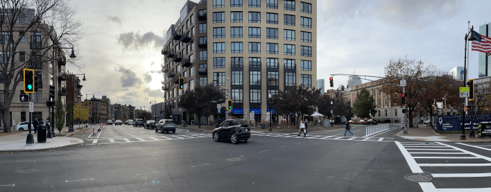 A panoramic image of two intersecting streets with a tall building in the middle of the image, on the opposite corner. Both streets include curbside bike lanes separated from traffic by cones or flexible-post bollards. Several pedestrians are crossing the street to the right on a crosswalk with the walk sign, while next to them a car drives through the intersection on the street to the left.