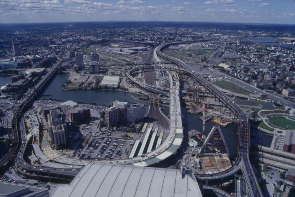 A historic color photograph of the North Station area in Boston looking north toward Cambridge. The train platforms of North Station are in the center, and above them are an under-construction highway viaduct. To the left of that bridge is a triangular concrete tower, which would later become the Zakim Bridge. To the right of that is an older double-decker highway, which was the previous route of I-93 through downtown Boston. The Charles River runs from left to lower right in the middle of the photo and I-93 meanders into the distant horizon at upper center.