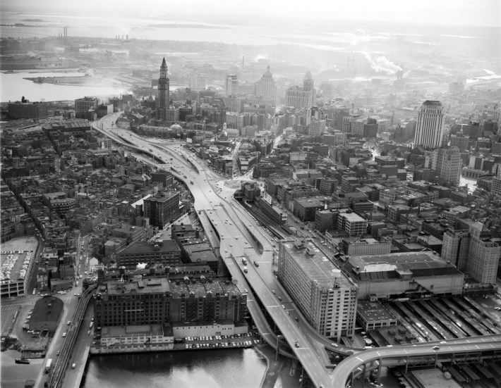 A black-and-white aerial view of downtown Boston in 1959. The Central Artery highway viaduct weaves through the center of the photo with on- and off-ramps descending to street level in a braided pattern. In the foreground are the Charles River (bottom edge) and the old Boston Garden (right lower corner). At upper left, near where the highway curves out of sight, is the Custom House Tower, then the tallest building in the city. South Boston and Dorchester Bay are visible in haze in the upper edge of the photo.