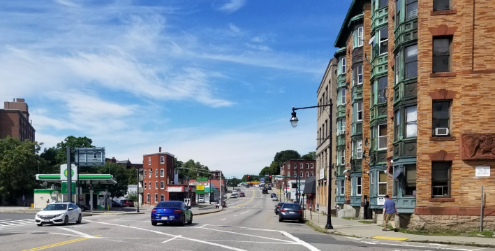A wide four-lane city street under a blue sky. On the right edge of the photo is a historic five-story brick apartment building with bay windows. On the left edge is a BP gas station. The wide roadway through the center of the photo only has a handful of cars on it, a few of which are parked at the curbside.