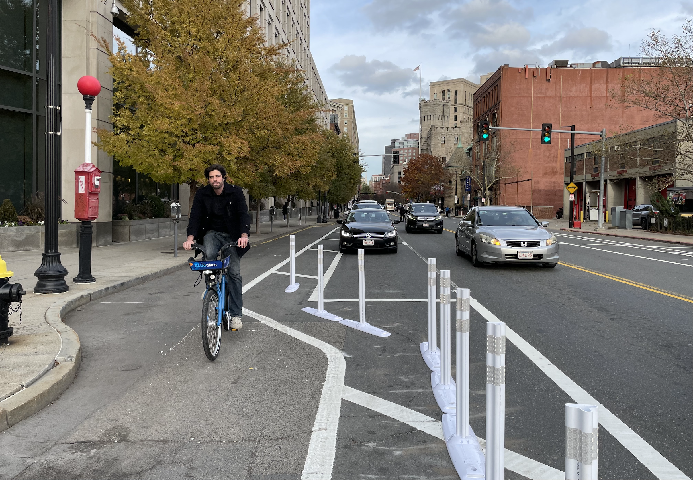 A man rides a Bluebike towards the camera in a separated bike lane that runs between the sidewalk (left) and a row of parked cars (center). A row of flexible-post bollards in the foreground divides the bike lane from moving traffic. The street is lined with multi-story buildings.