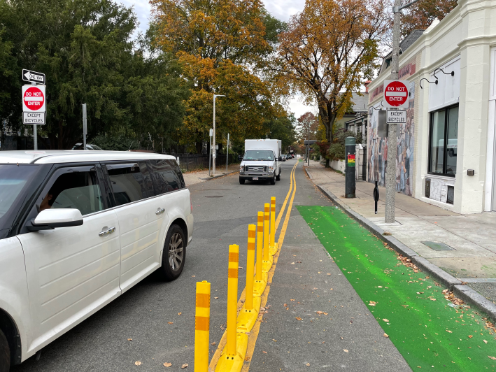 A green-painted bike lane separated from the rest of the street by a row of yellow flexible-post bollards. On either side of the street are two "Do Not Enter – Except Bicycles" signs. Beyond the yellow bollards is a pair of yellow-striped lines that divide the bike lane from motor vehicle traffic coming from the opposite direction. On the right is a one-story commercial building with a mural. Trees with yellowing leaves line the street in the distance.
