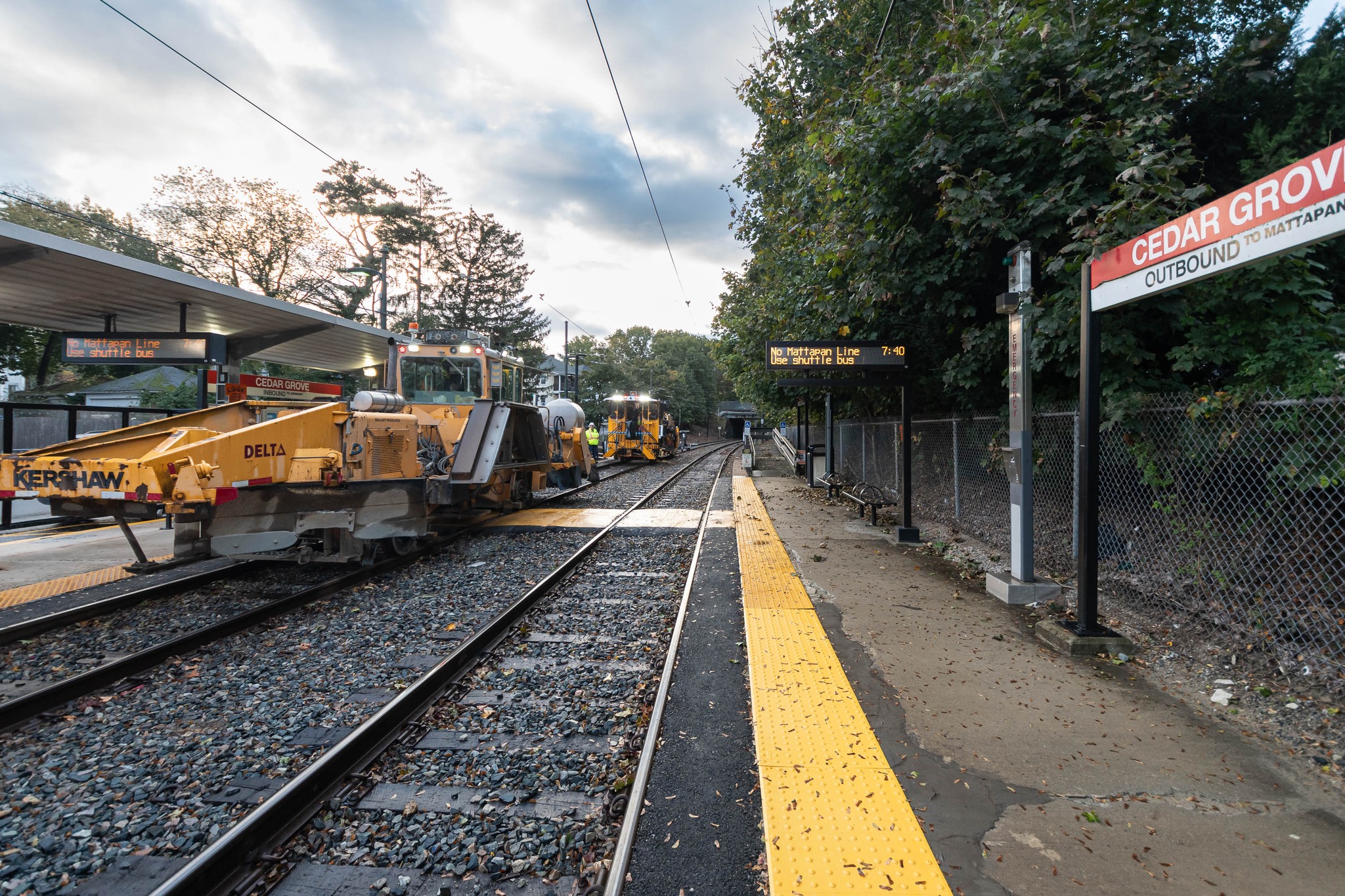 A yellow maintenance train sits on the tracks next to a platform of a train station. A red-and-white sign on the right platform, which is adjacent to a second empty track, shows that this station is Cedar Grove on the MBTA red line.