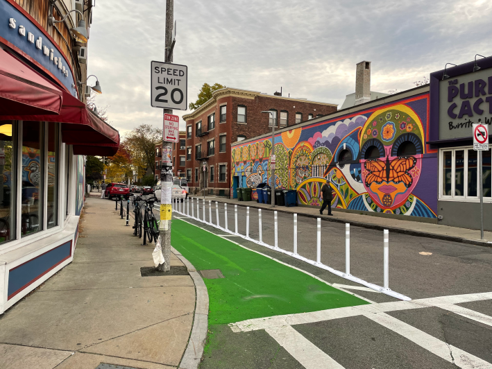A green-painted bike lane separated from the rest of the street by a row of flexible-post bollards next to a "speed limit 20" sign on the curb. Several bikes are parked in bike racks in front of a sandwich shop to the left. On the opposite side of the street is a brightly-painted mural on a one-story building next to a sign for "The Purple Cactus Burritos and Wraps"