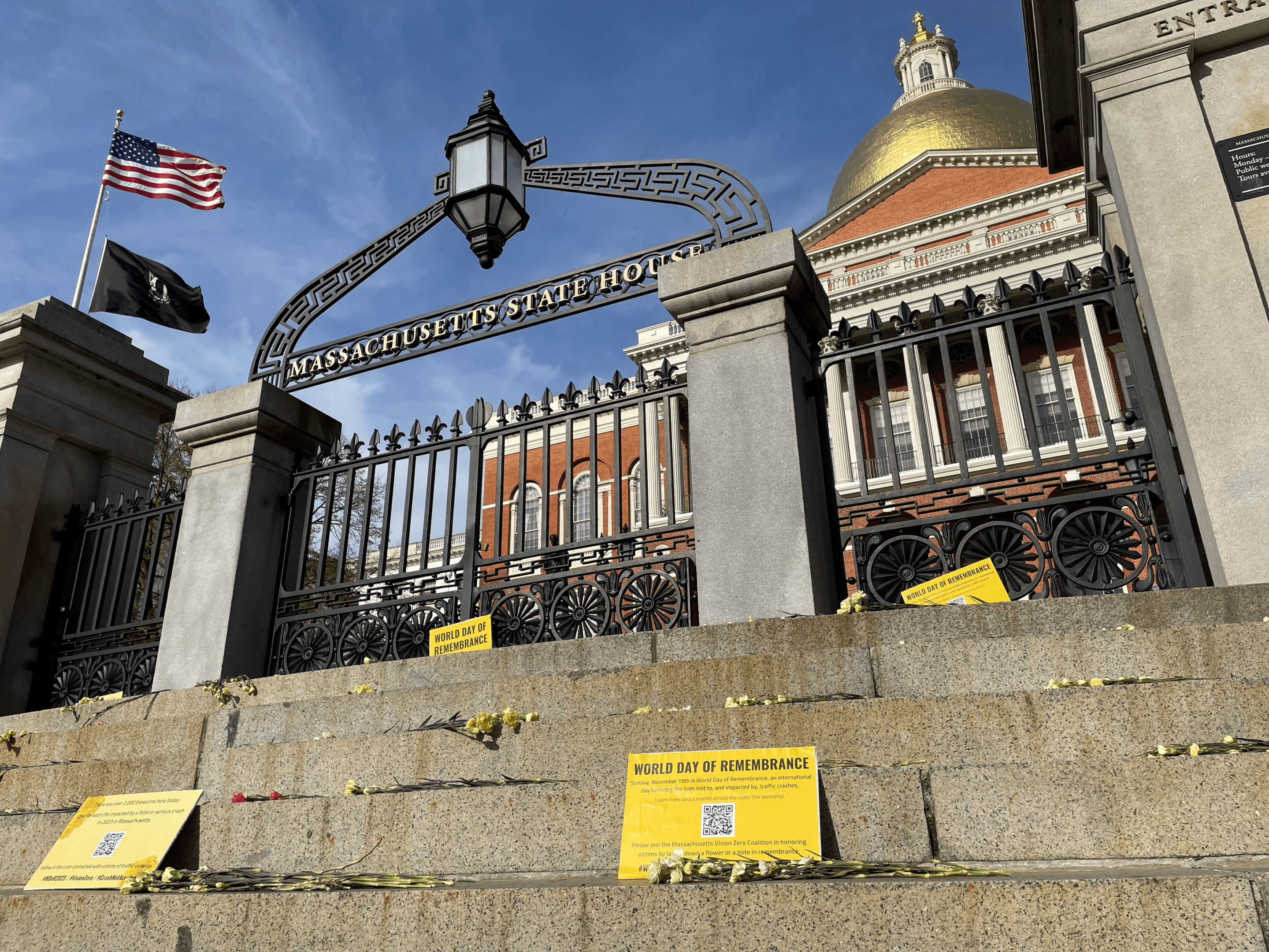 Hundreds of yellow flowers are scattered on the steps of the Massachusetts State House, with the gold-plated dome of the State House visible at upper right. Signs proclaim the flowers are in honor of the victims of traffic violence for the World Day of Remembrance.