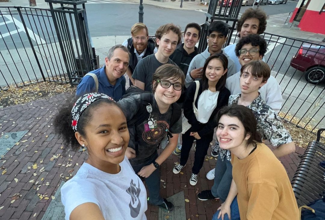A group of high school students and their chaperones pose for a selfie in front of the wrought-iron gate of a city park.