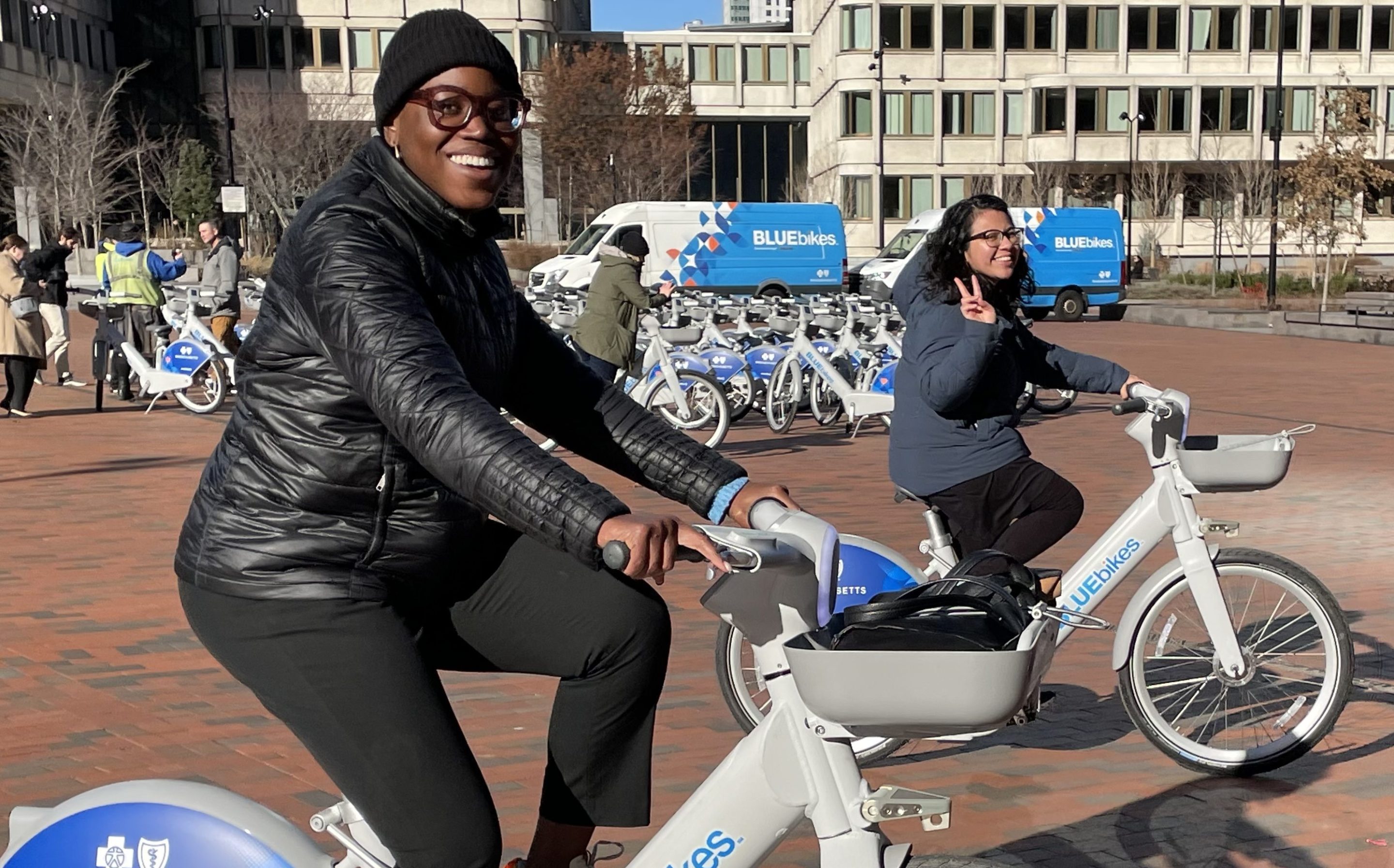 A young black woman with glasses and a black puffy winter coat and a young Hispanic woman in a blue winter coat holding up two fingers in a peace sign pedal on new electric Bluebikes through City Hall Plaza. Behind them are rows of more new bikes and office buildings in the distance.