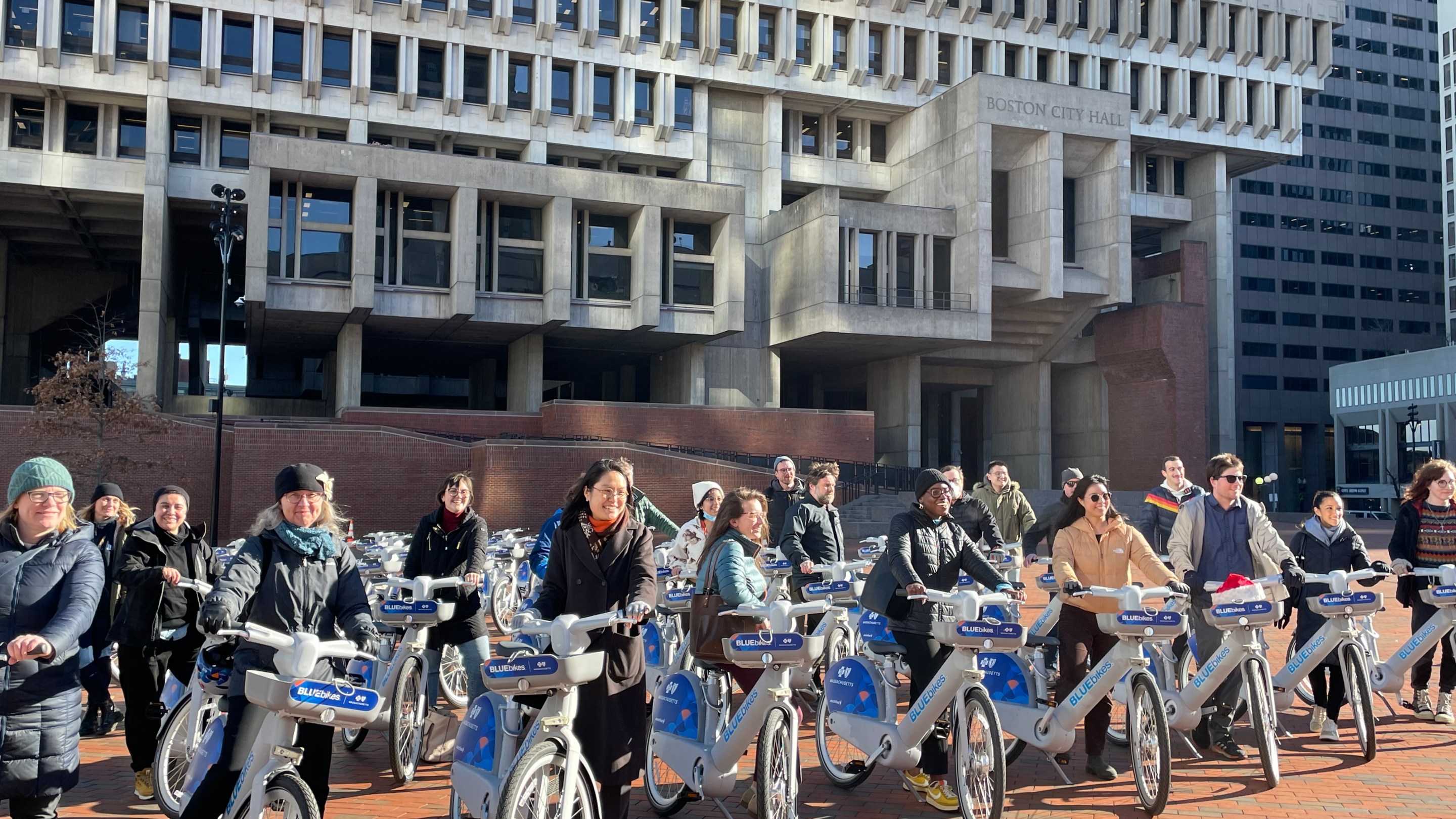 A crowd of people stand with new electric bikes in front of Boston City Hall