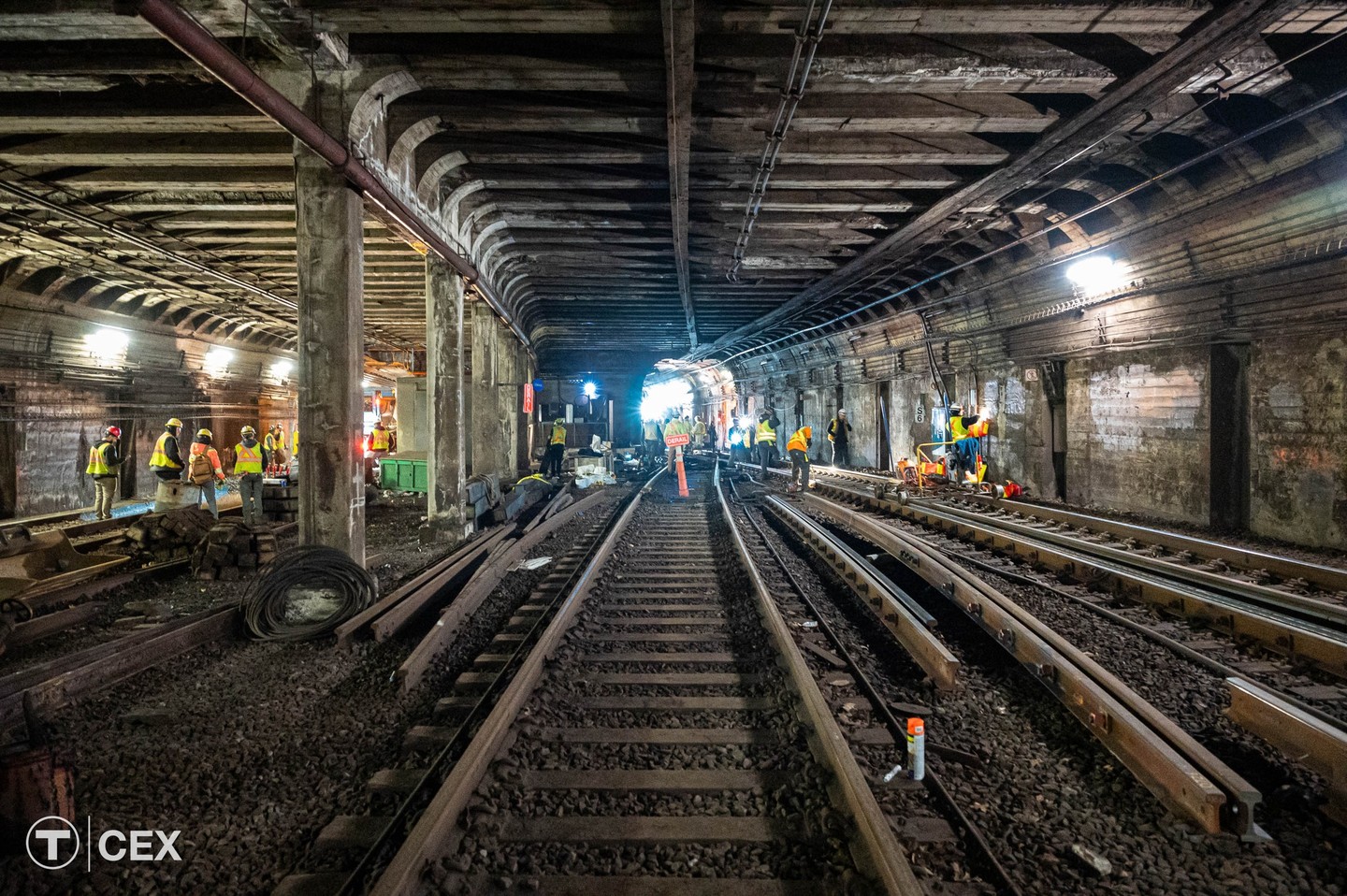 Workers in neon yellow vests work among rail tracks in a wide tunnel under bright electric lights.