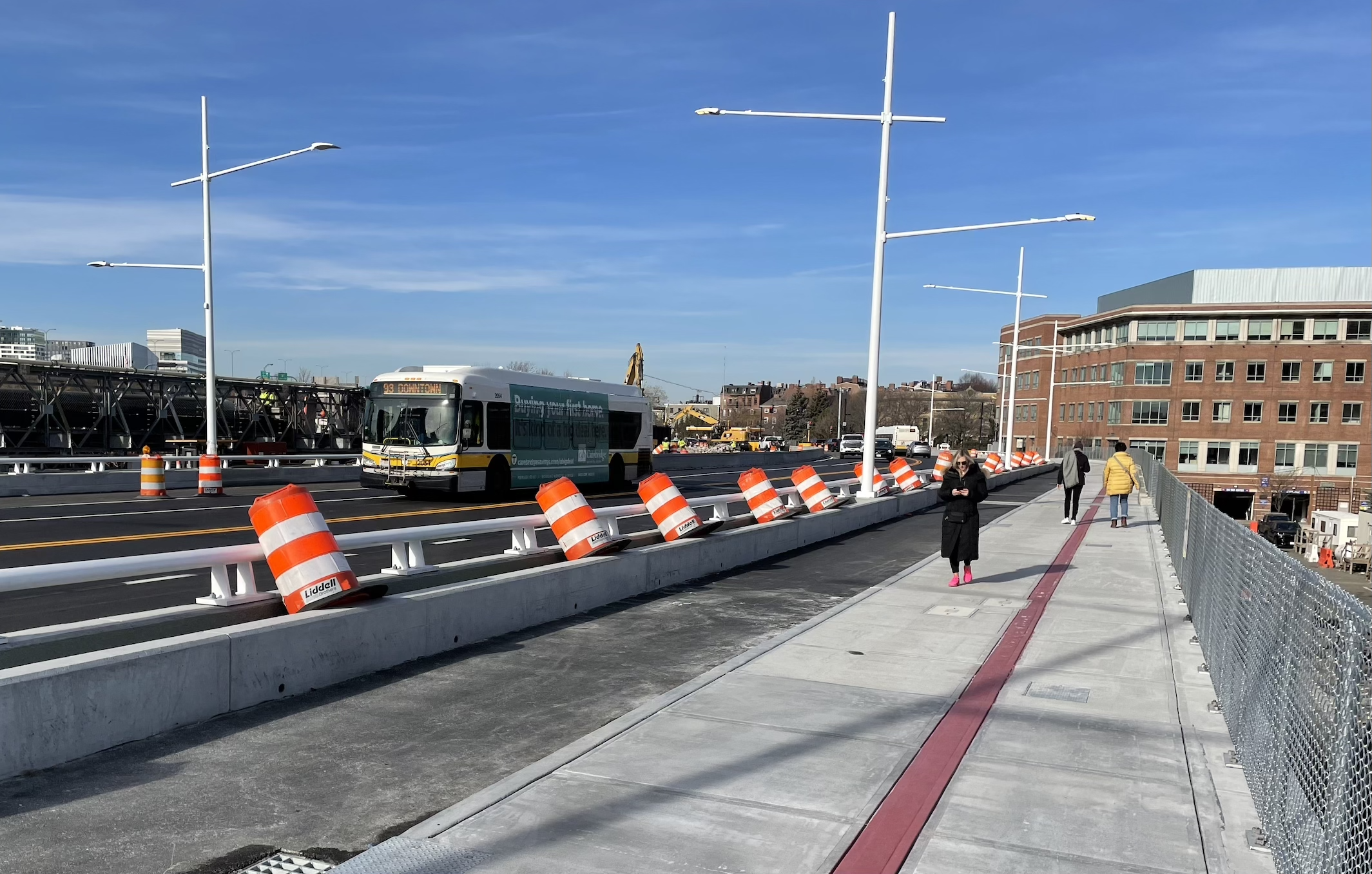 An MBTA 93 bus drives across a new bridge while several pedestrians use the wide sidewalk along the right edge of the photo.