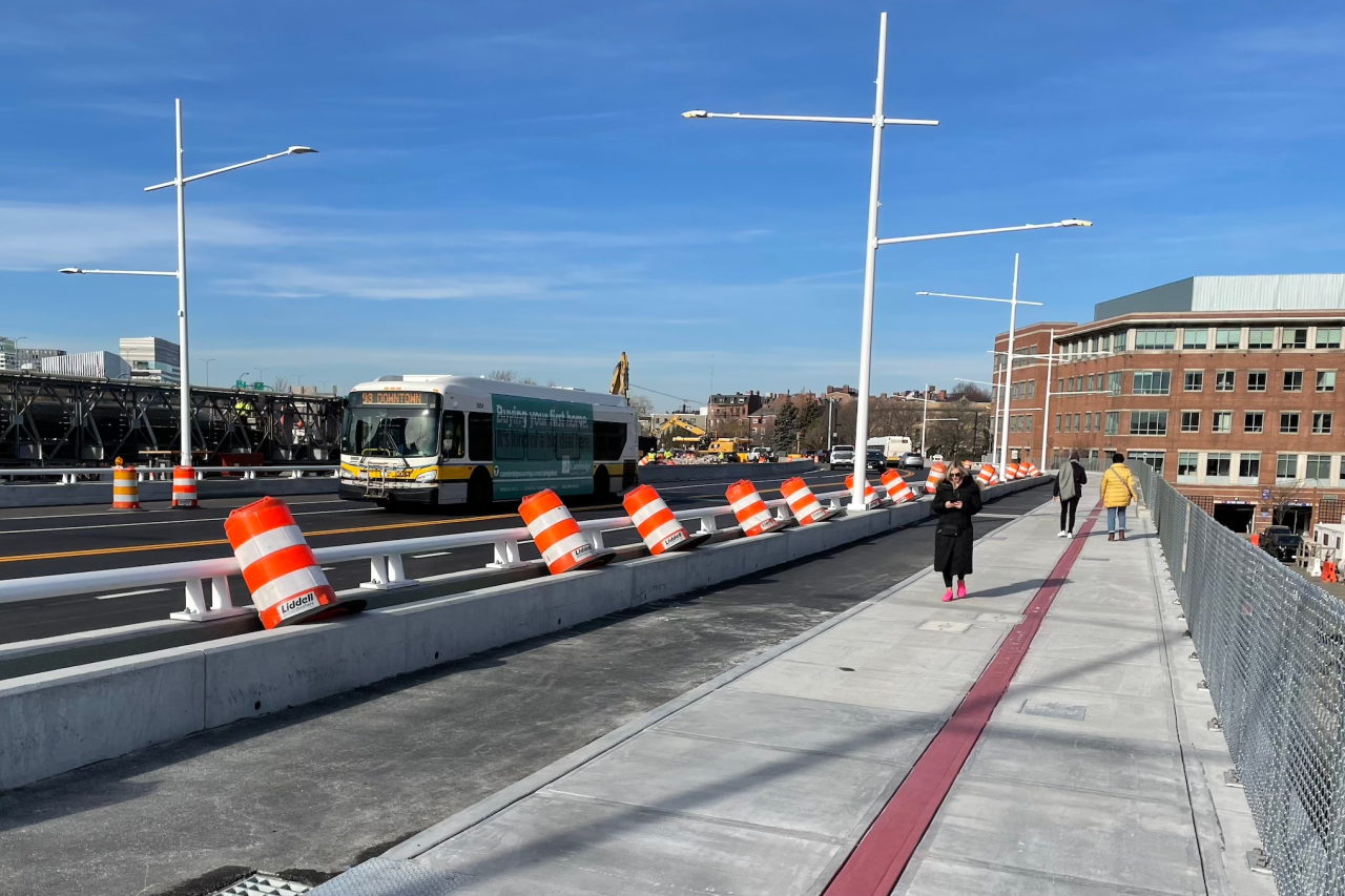 An MBTA 93 bus drives across a new bridge while several pedestrians use the wide sidewalk along the right edge of the photo.